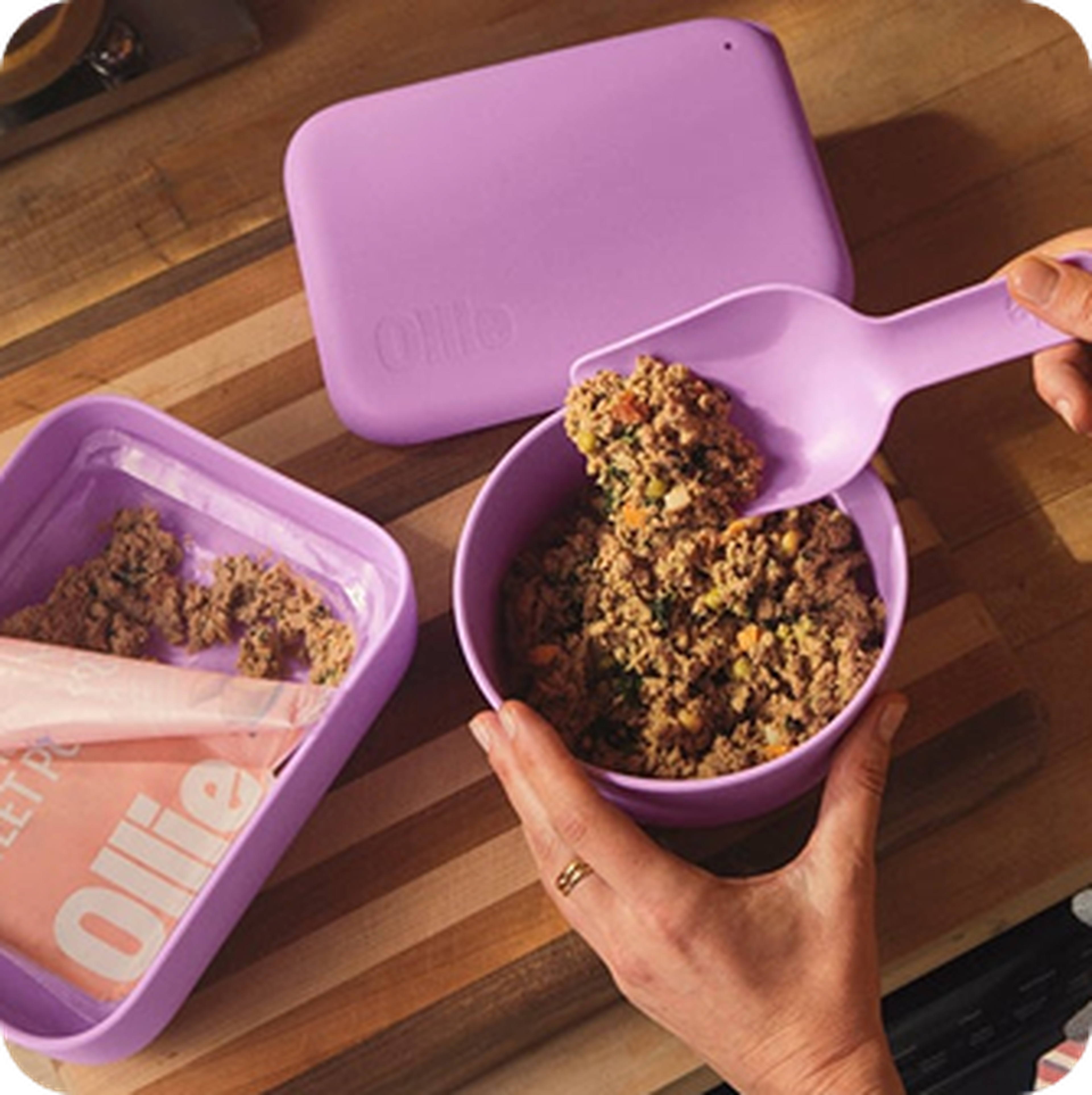 Hands scooping pet food from a purple container into a purple bowl on a wooden countertop. Another container is open beside them.