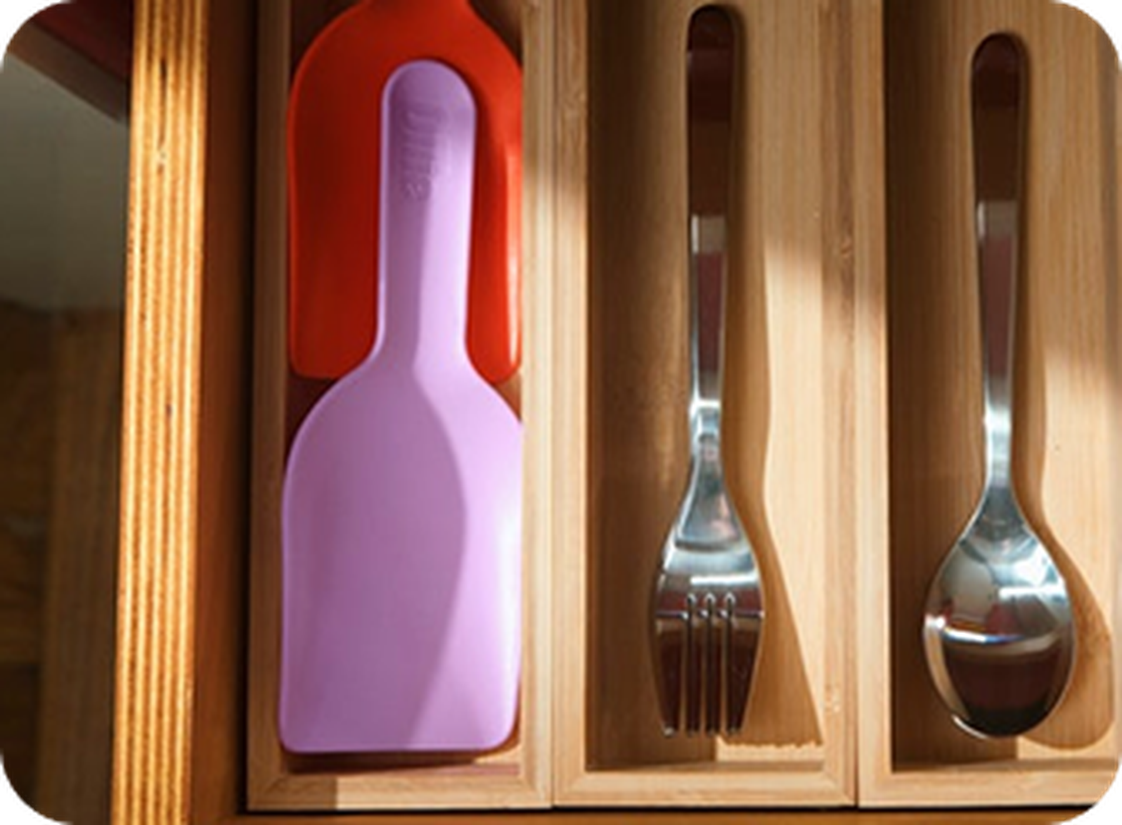 Wooden cutlery drawer with sections holding red and purple spatulas, forks, and spoons. Sunlight casts shadows on the cutlery.