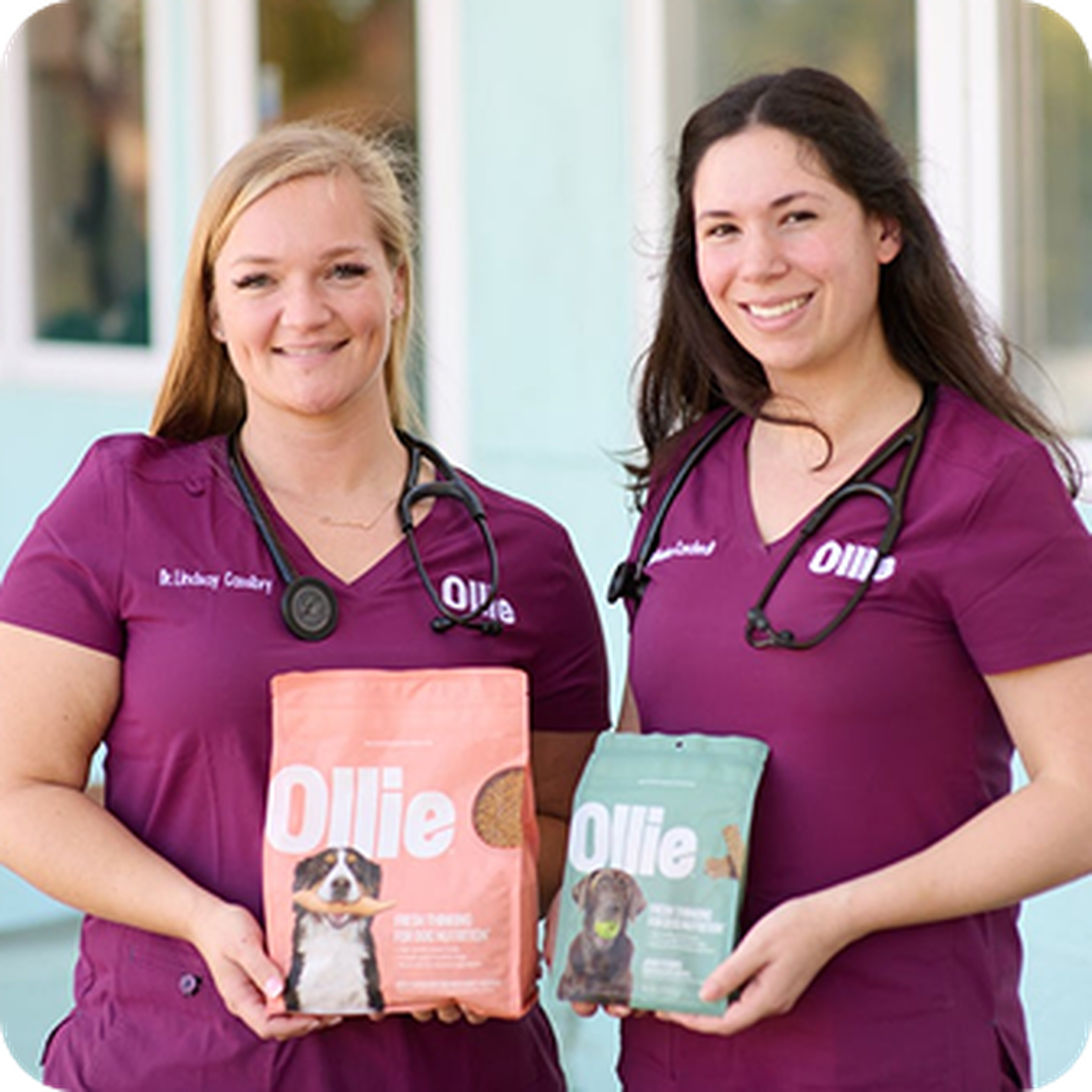 Two women in purple scrubs, wearing stethoscopes, smiling and holding bags of dog food labeled "Ollie."