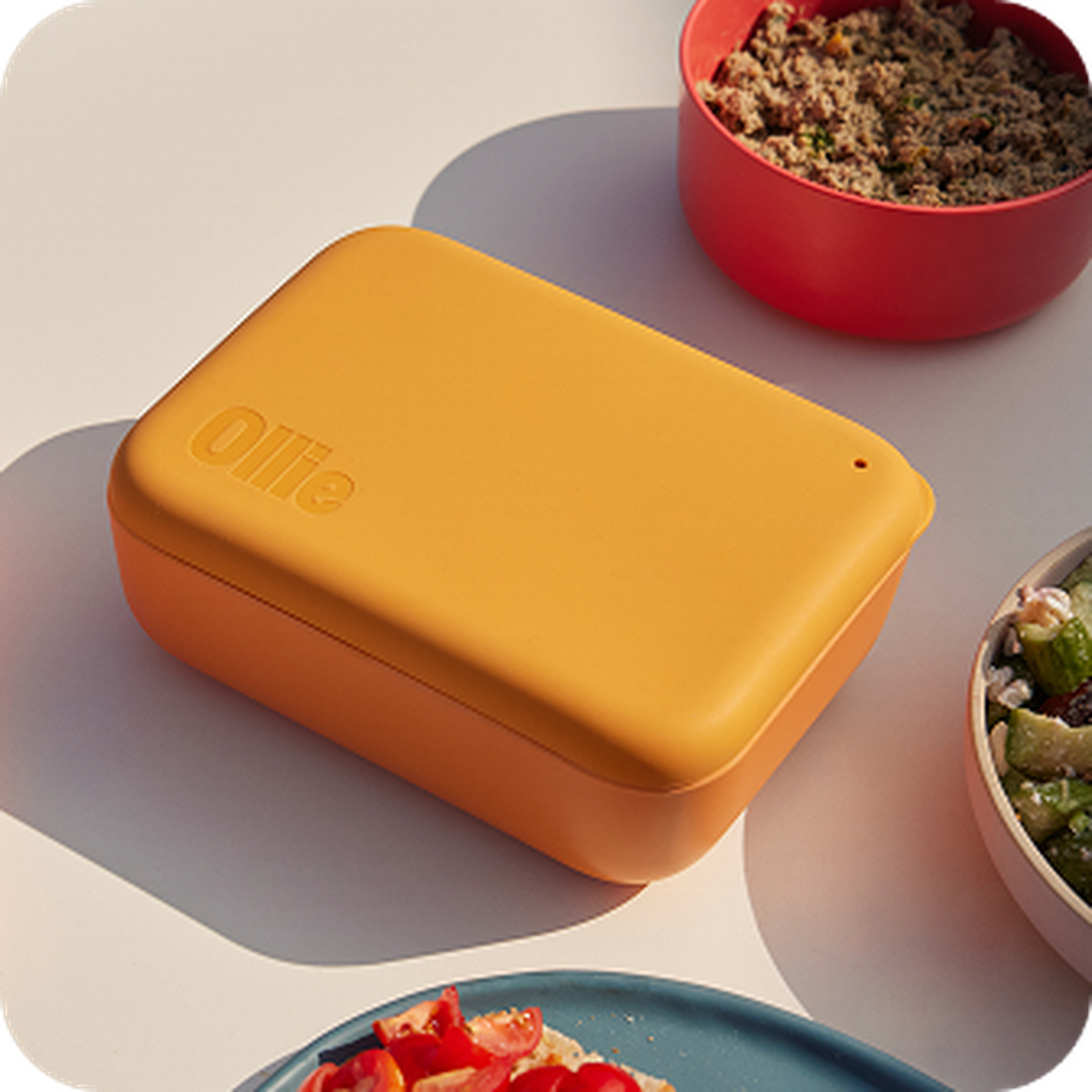 Yellow rectangular container with "Ollie" embossed on the lid, surrounded by bowls of food on a white surface.