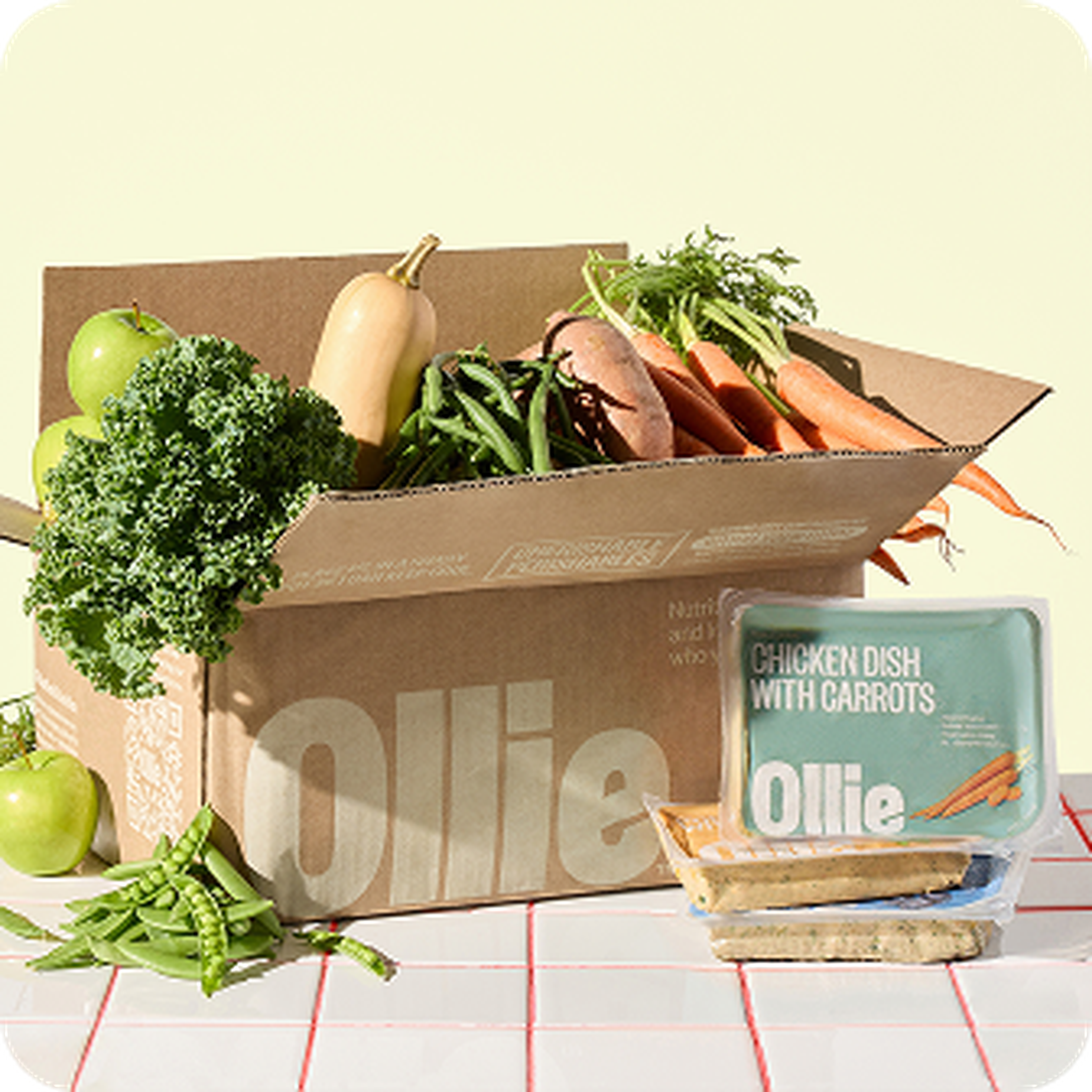 Cardboard box with fresh vegetables: kale, squash, beans, carrots, sweet potatoes, apples. Nearby, Ollie-packed chicken dish with carrots.
