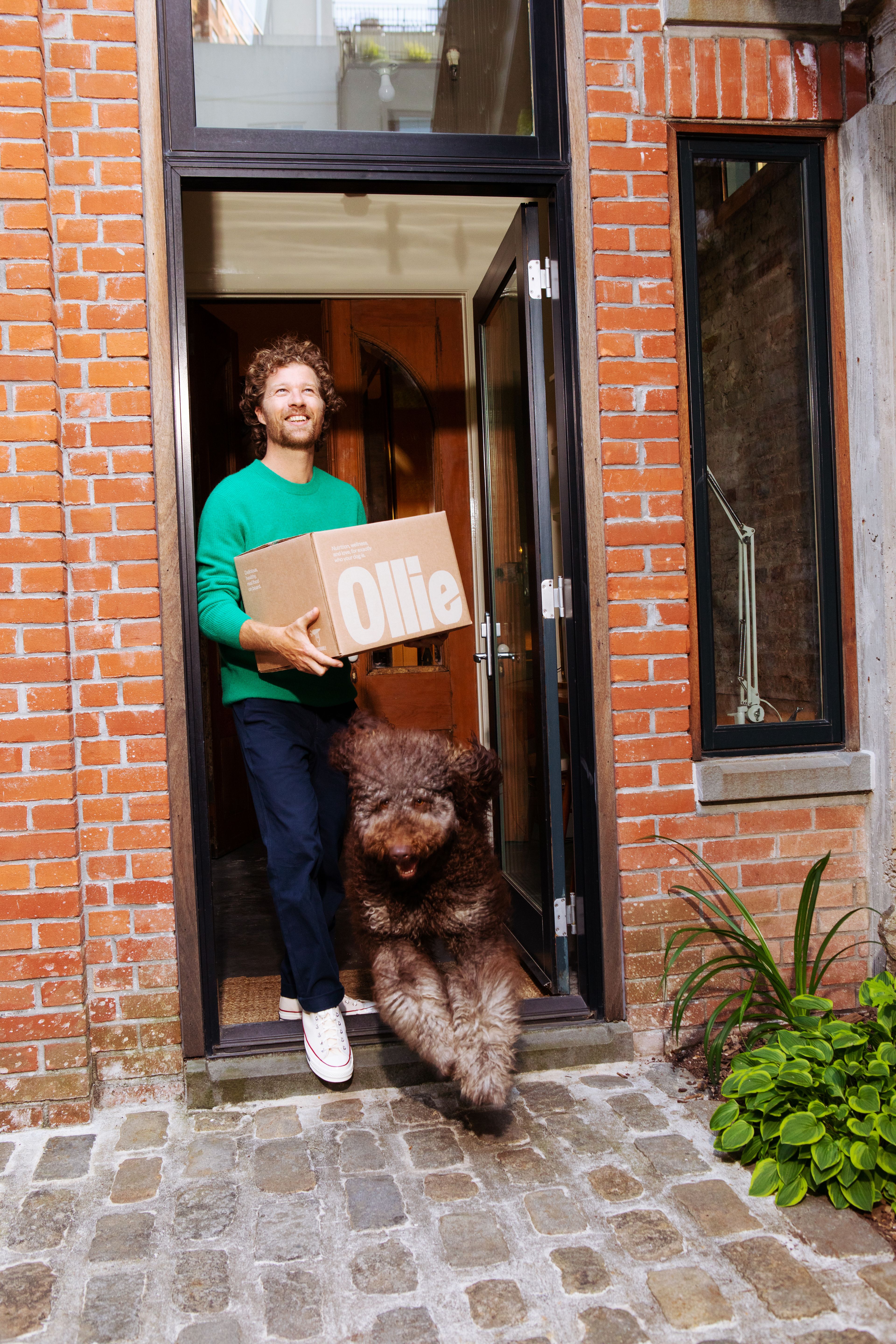 man holding Ollie box in doorway while brown fluffy dog jumps out