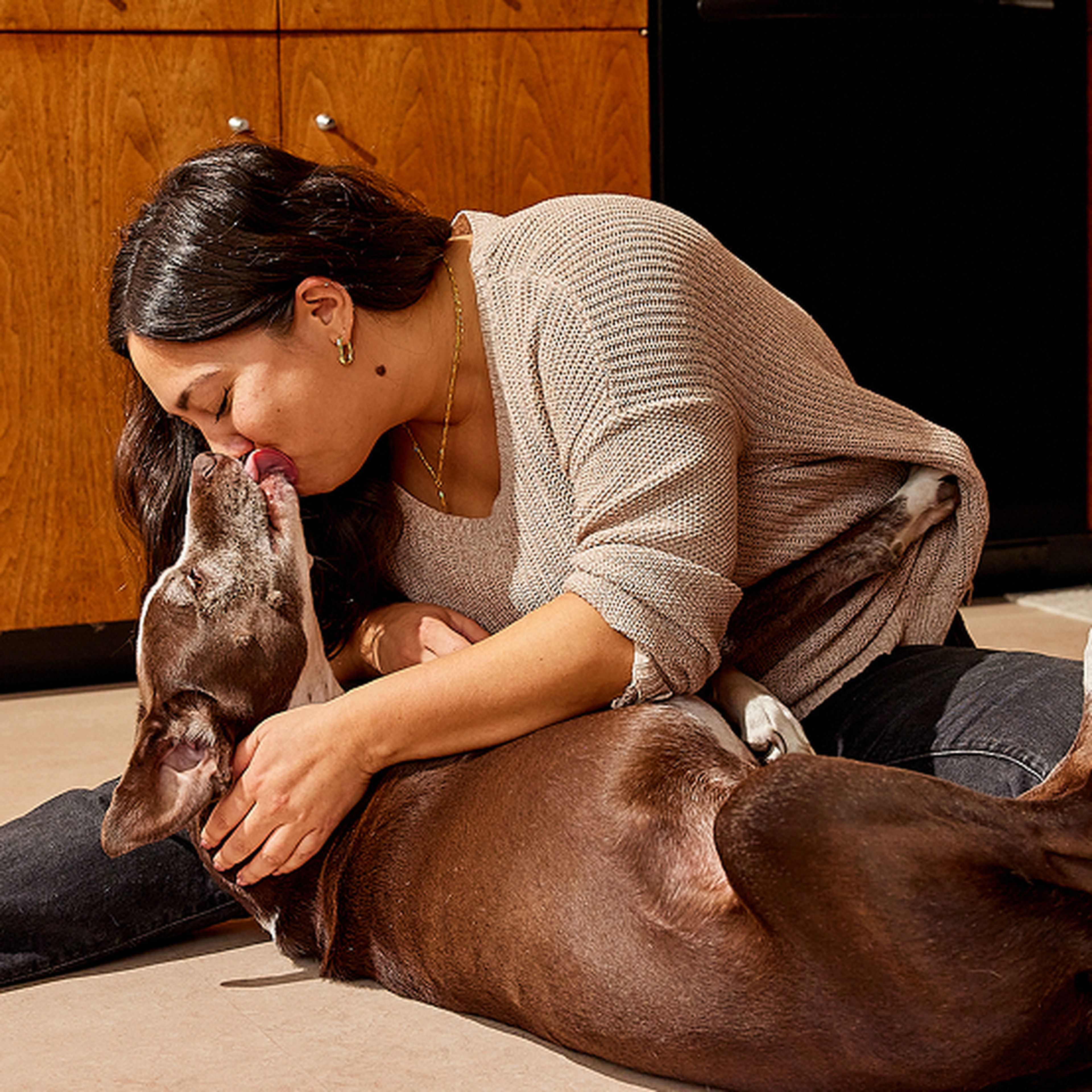 Woman lovingly embraces and kisses a brown dog on the floor in a cozy kitchen setting.