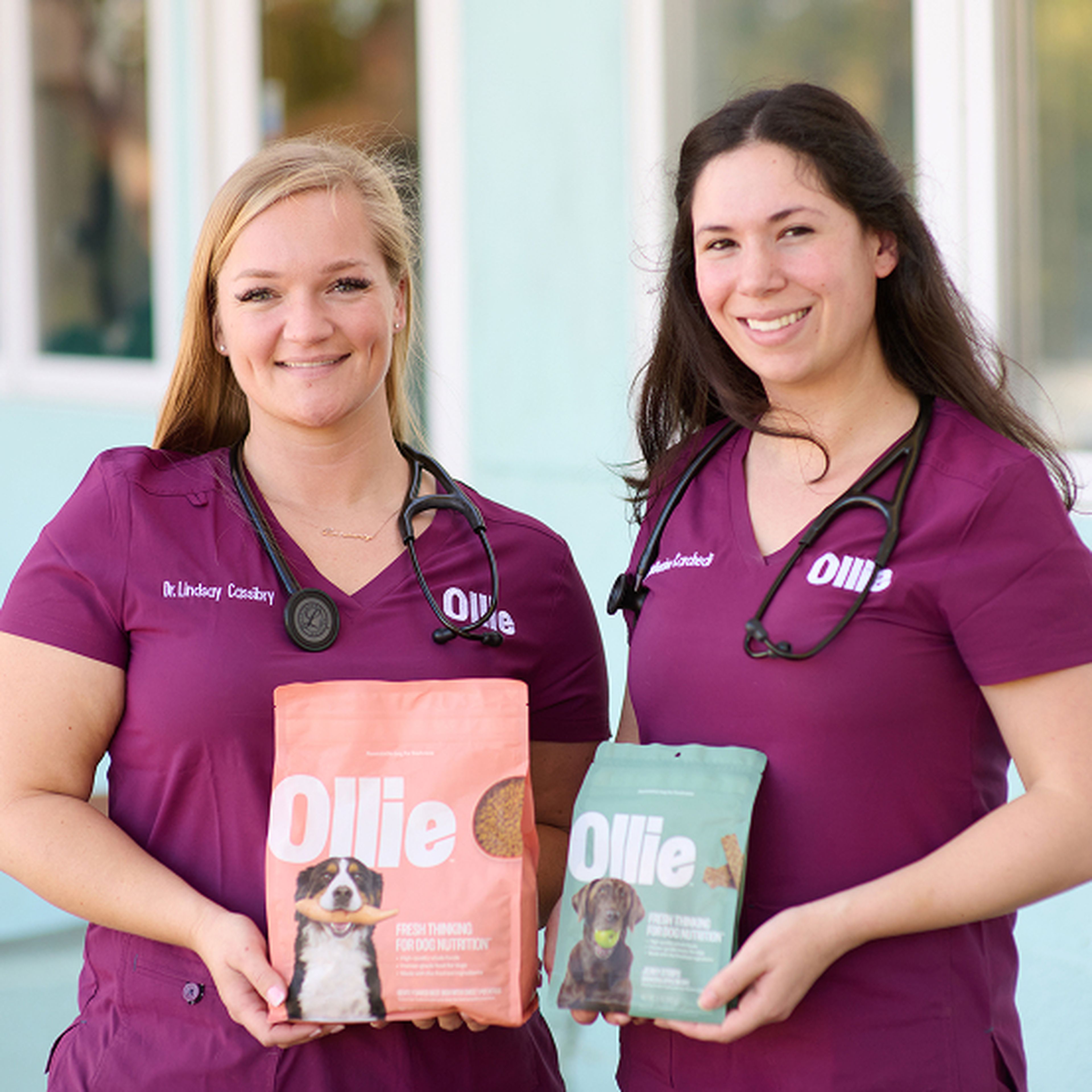 Two women in purple scrubs with stethoscopes hold Ollie dog food packages, standing outside a building.
