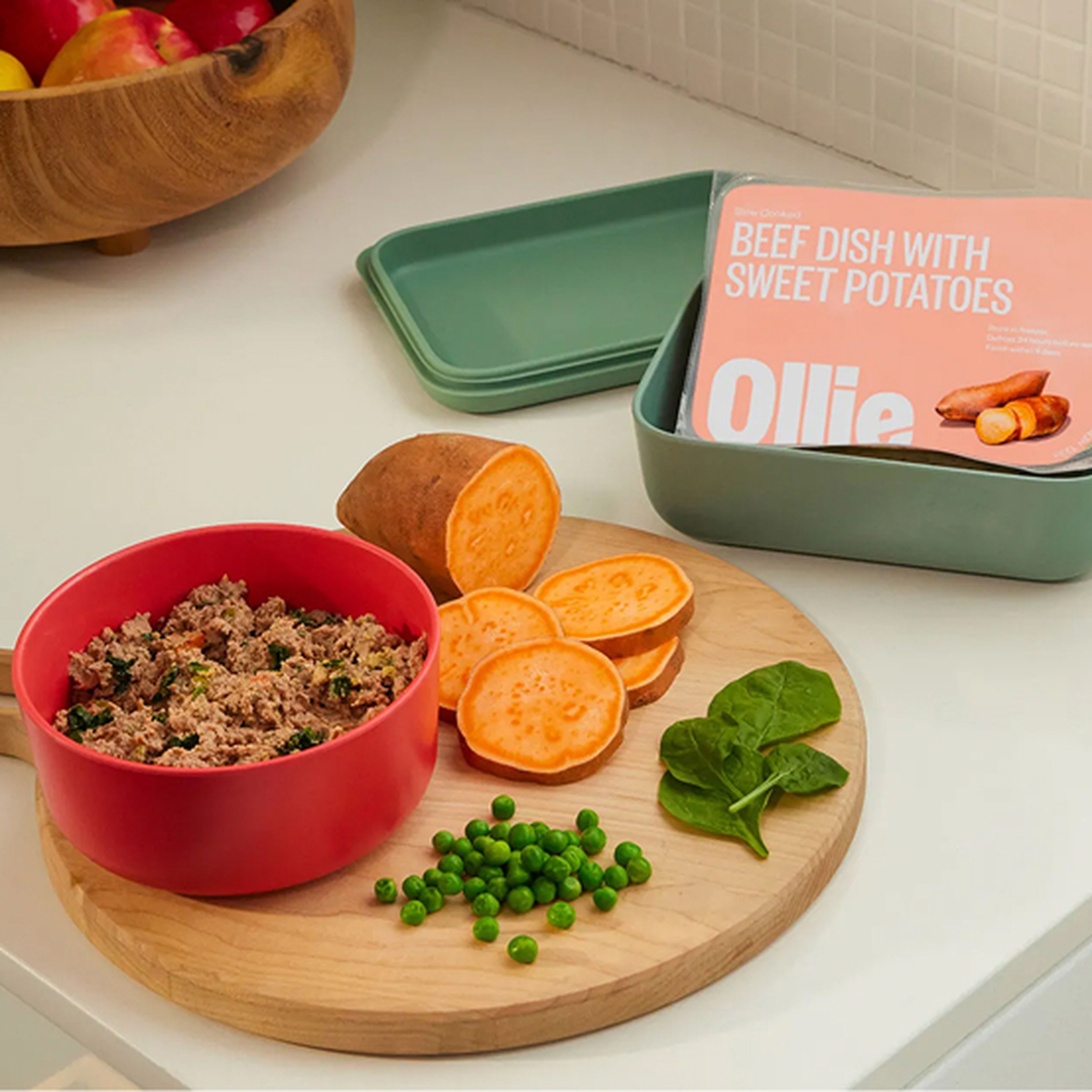 A red bowl with beef, peas, and spinach next to sliced sweet potatoes on a cutting board. Ollie dog food packaging is in the background.