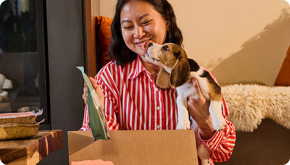 Woman in a red striped shirt smiling as a puppy licks her face, sitting next to an open cardboard box with pet supplies.