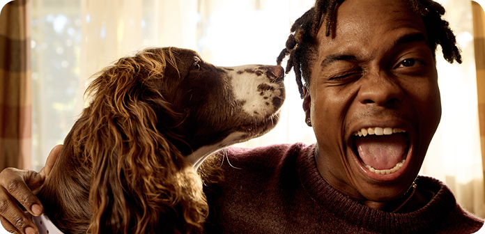 Man winks playfully while hugging a dog. The dog faces him, appearing to sniff his cheek. Both seem joyful and energetic indoors.