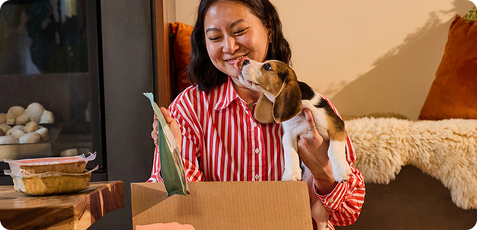 Woman in a red striped shirt smiling at a beagle puppy while unpacking a cardboard box in a cozy room.