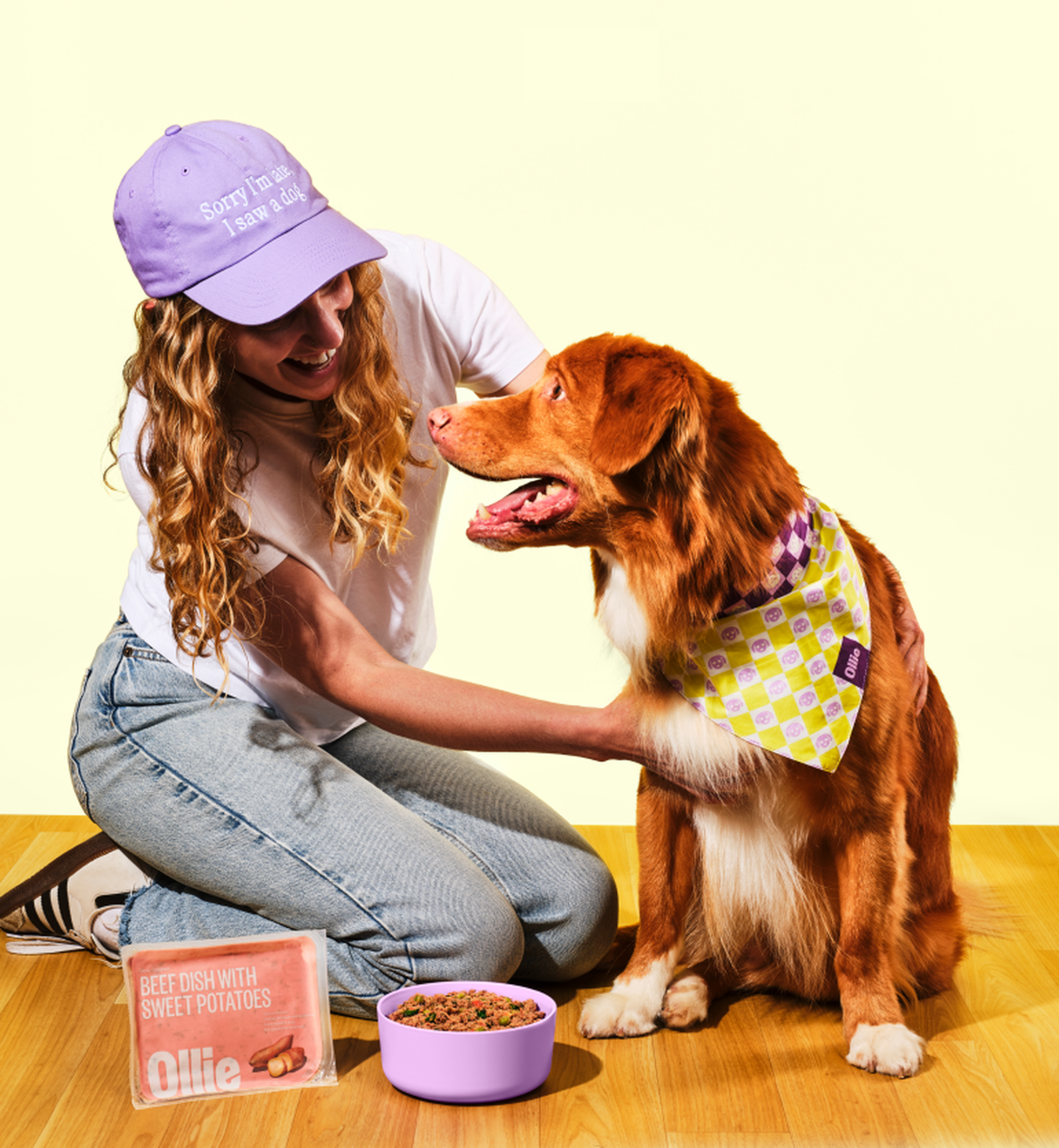 A person with curly hair and a purple cap smiles at a dog wearing a bandana. Nearby are a bowl of dog food and a packaged meal.