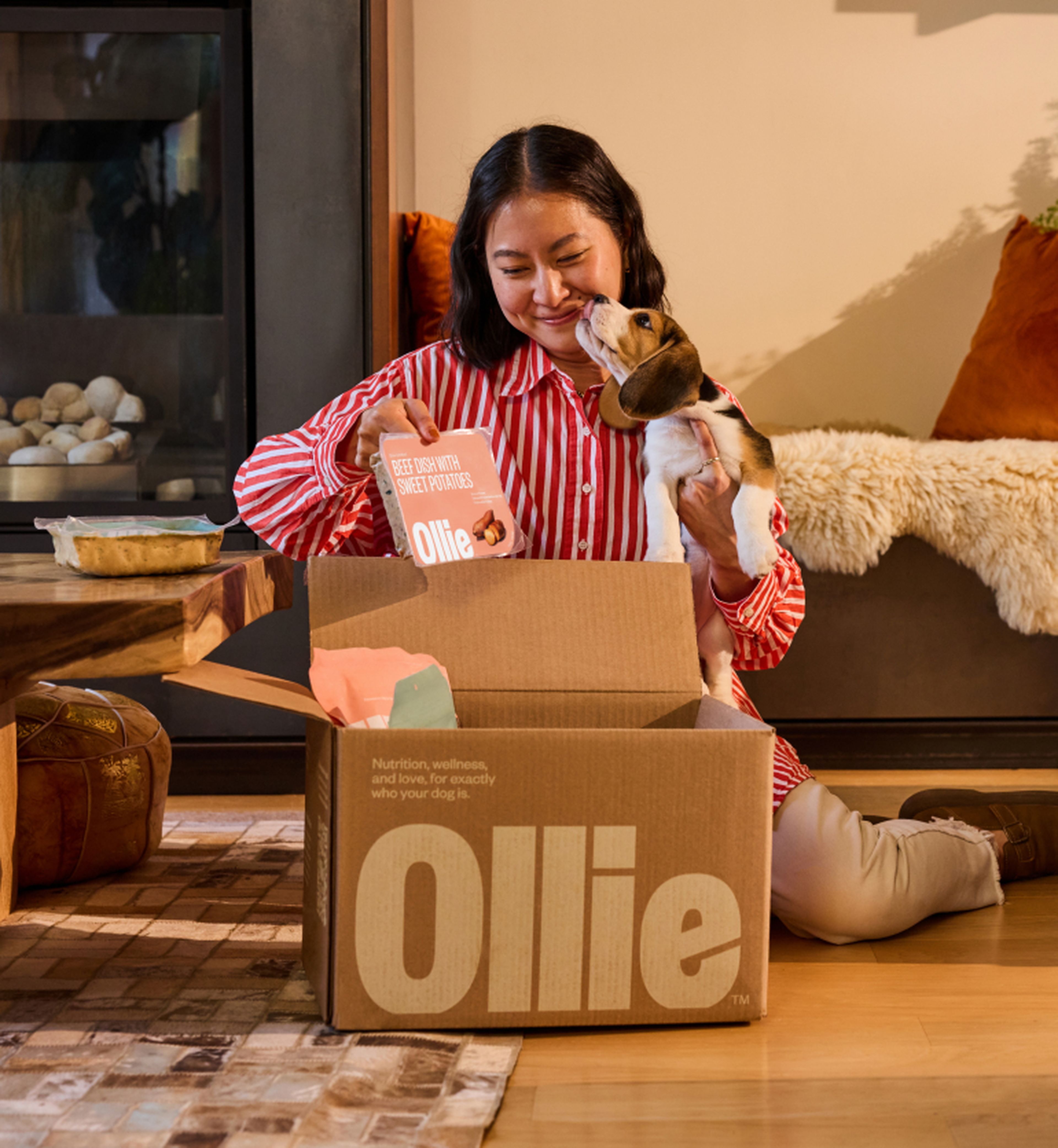 Woman sits on the floor opening an Ollie pet food box, smiling at a small dog in her arms in a warmly lit living room.