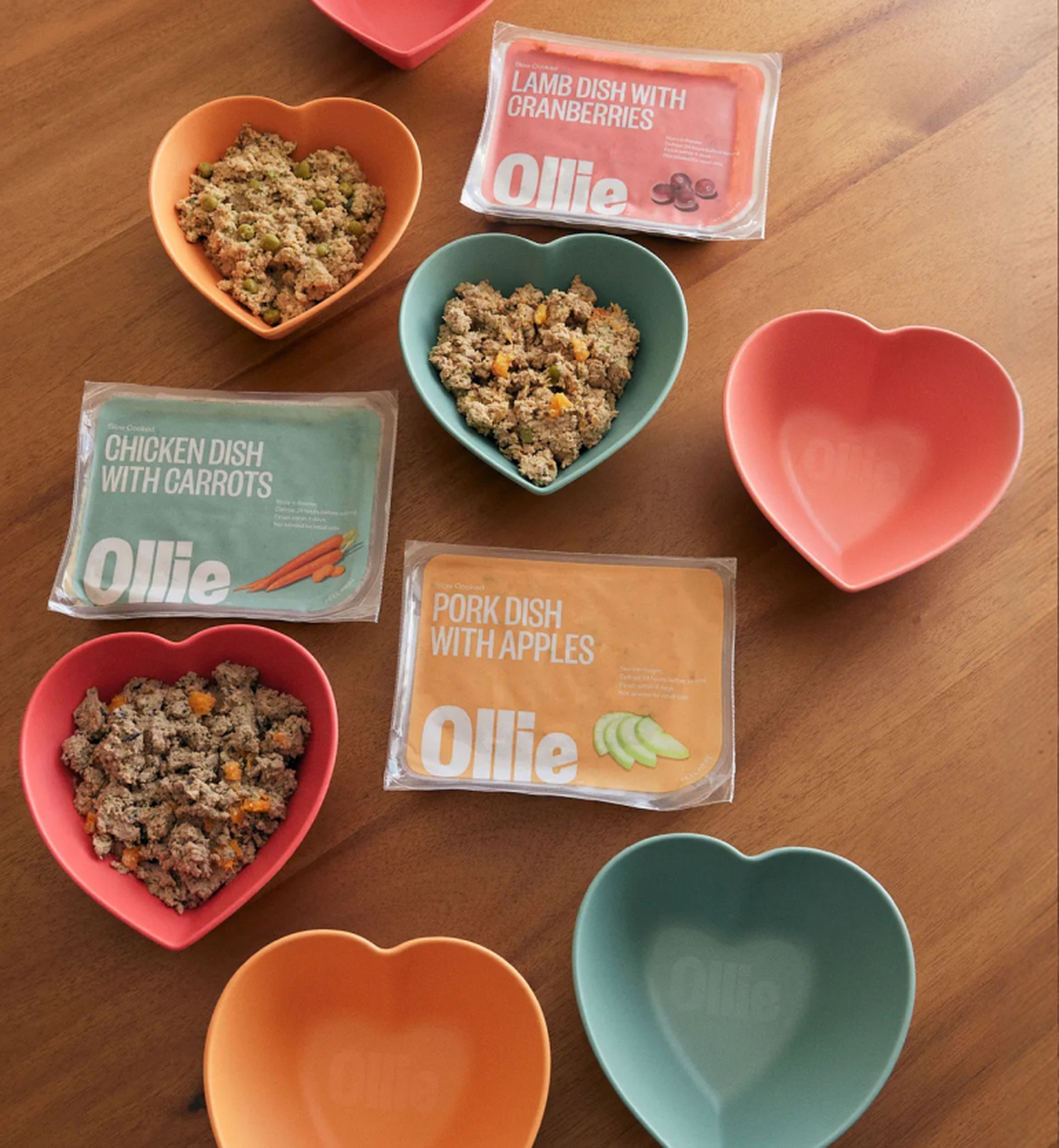 Heart-shaped bowls and packaged meals for dogs are arranged on a wooden table, including chicken, pork, and lamb dishes from Ollie.