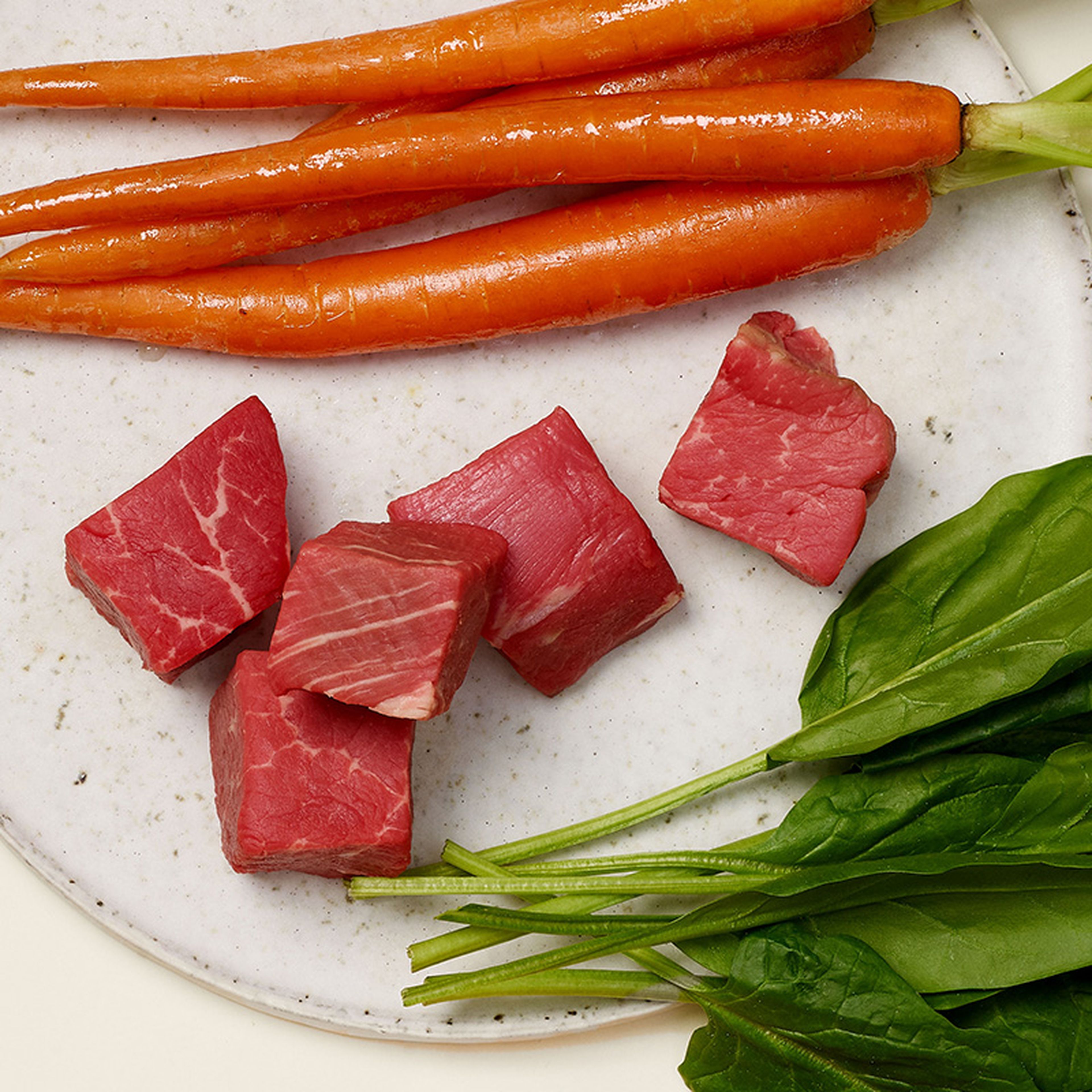 Plate with raw beef cubes, fresh carrots, and spinach leaves on a light background.