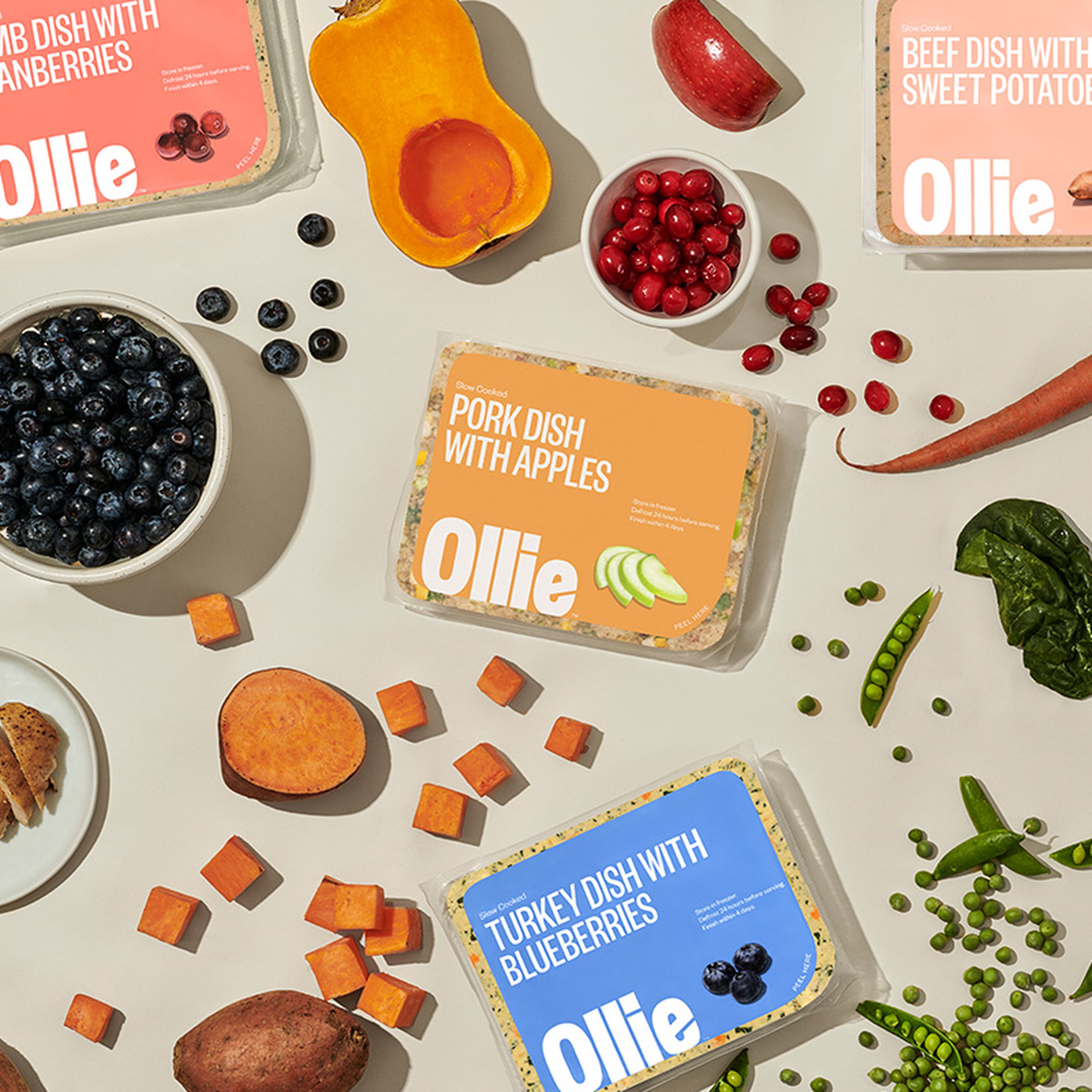 Assorted Ollie dog food packages surrounded by fresh ingredients like sweet potatoes, blueberries, peas, and cranberries on a light surface.
