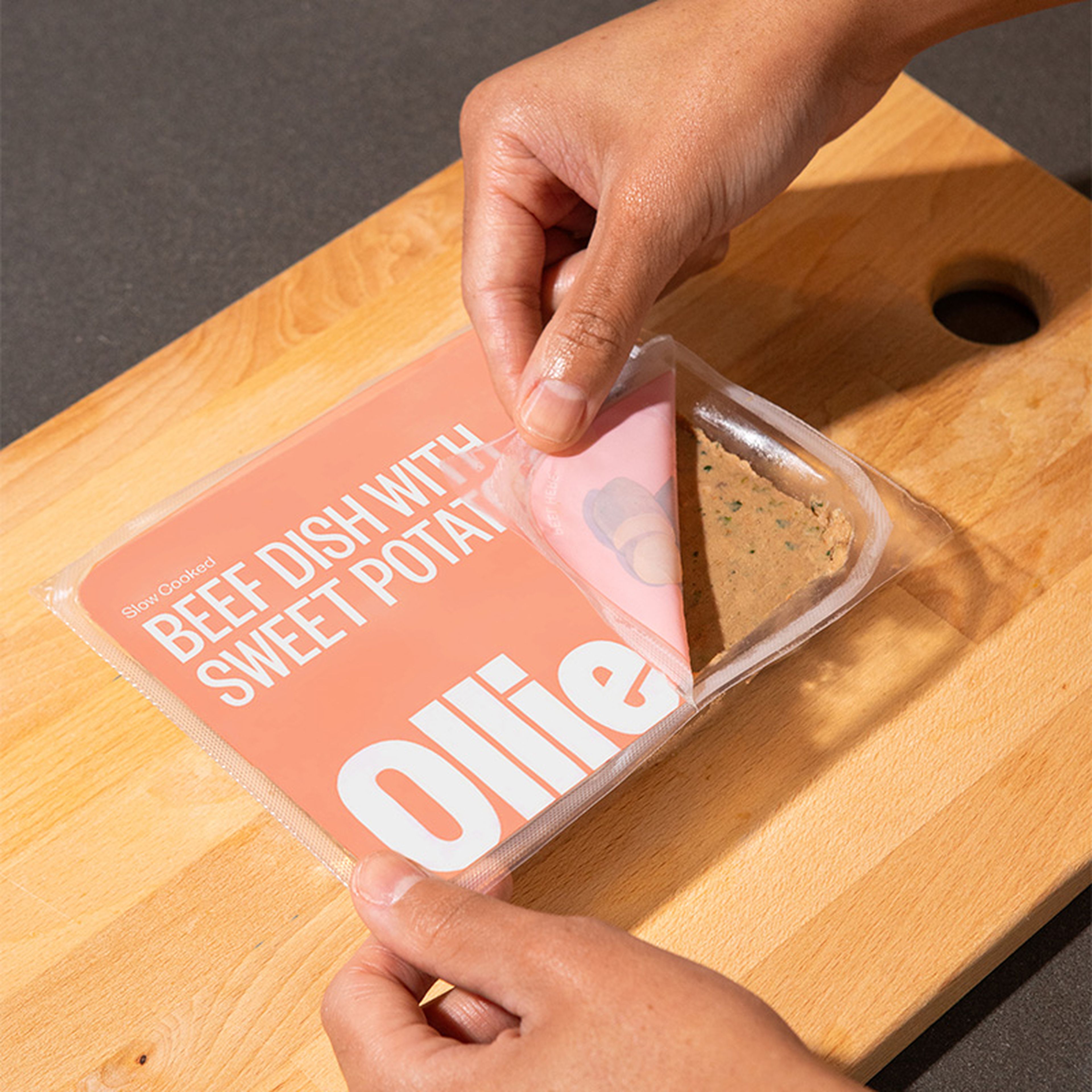 Hands opening a packaged meal labeled "Beef Dish with Sweet Potato" on a wooden cutting board.