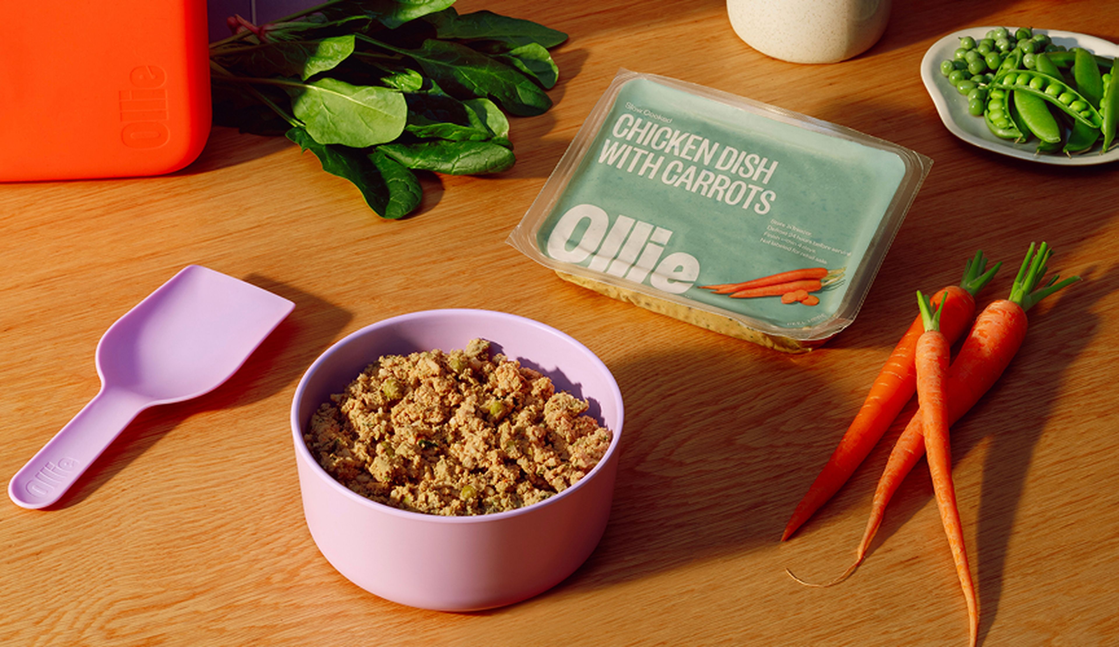 Dog food bowl filled with chicken and carrots, a pink scoop, fresh carrots, spinach, and peas on a wooden table, with Ollie packaging.
