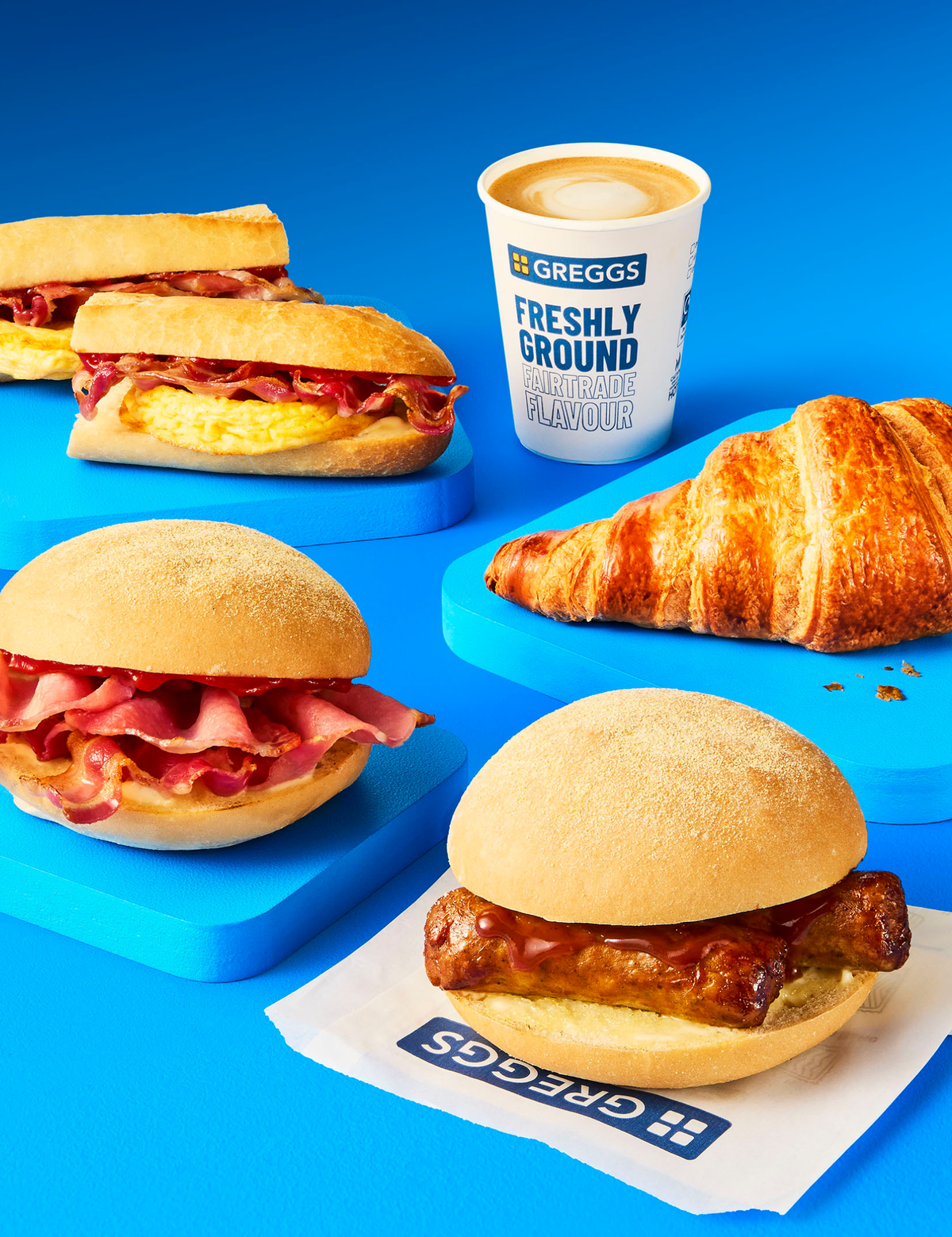A selection of breakfast goodies on a blue background - a bacon breakfast roll, a sausage breakfast roll, a latte