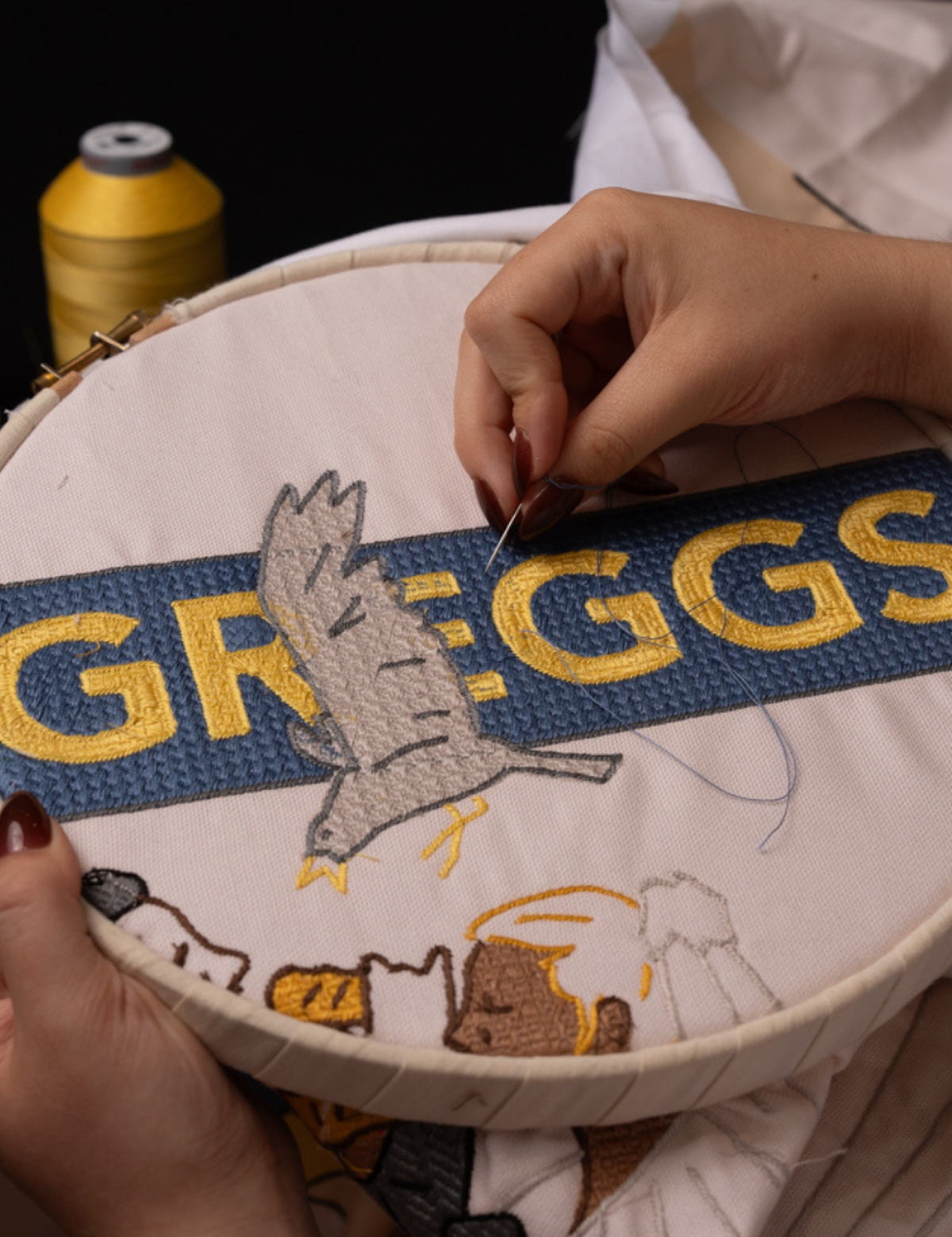 A scene of a Greggs logo and seagull, stitched onto cotton fabric in golden and blue thread