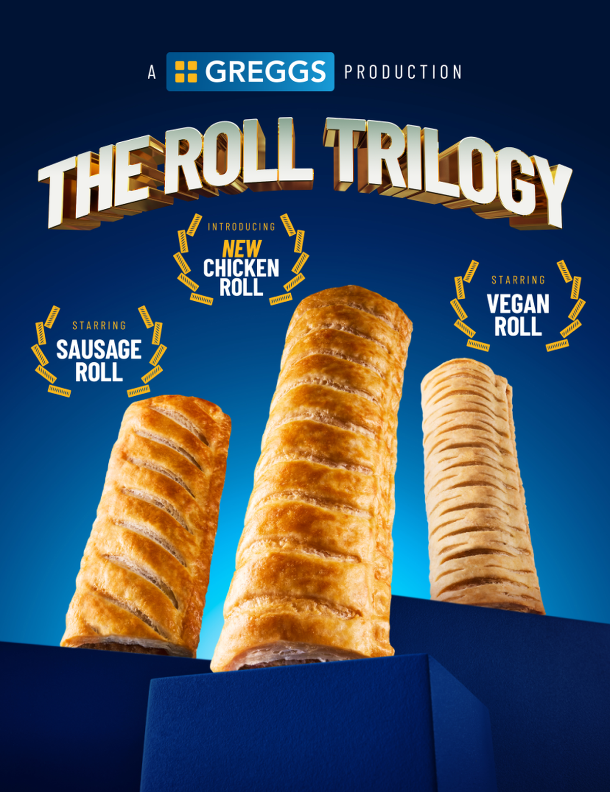 Three rolls on podiums, illuminated by a spotlight. Beside them, "NEW Chicken Roll", "Vegan Roll" and "Sausage Roll" are written in Sausage Roll styled rosettes, and "The Roll Trilogy" is written in an epic golden font