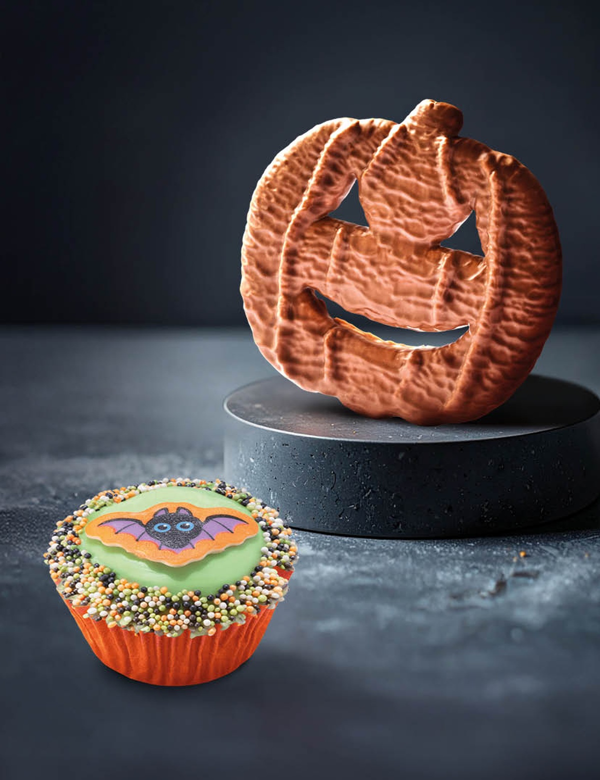 A Halloween-themed cupcake with a bat design and sprinkles next to a biscuit in the shape of a carved pumpkin on a dark pedestal.