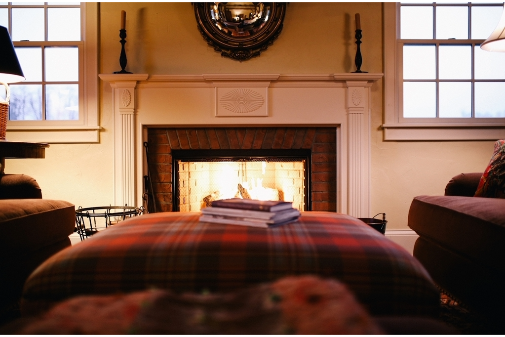 Cozy living room with a lit fireplace, flanked by windows. Plaid ottoman and couches create a warm, inviting atmosphere.