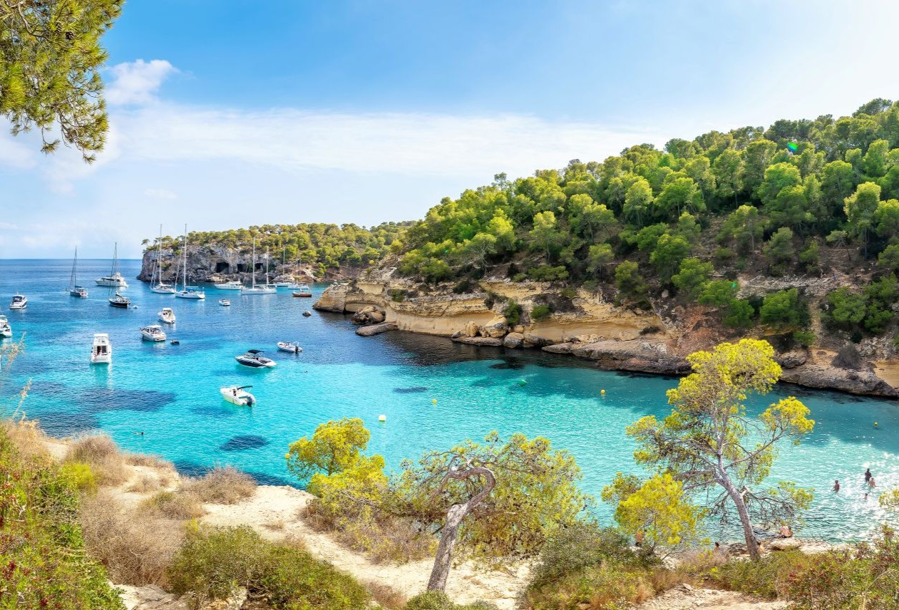 Scenic view of a turquoise bay with boats, surrounded by lush green hills and rocky cliffs under a clear blue sky.
