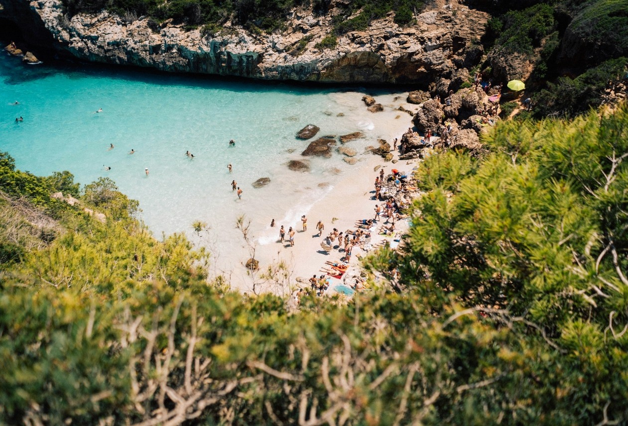 Aerial view of a small beach with turquoise water, surrounded by rocky cliffs and lush greenery. People sunbathe and swim.