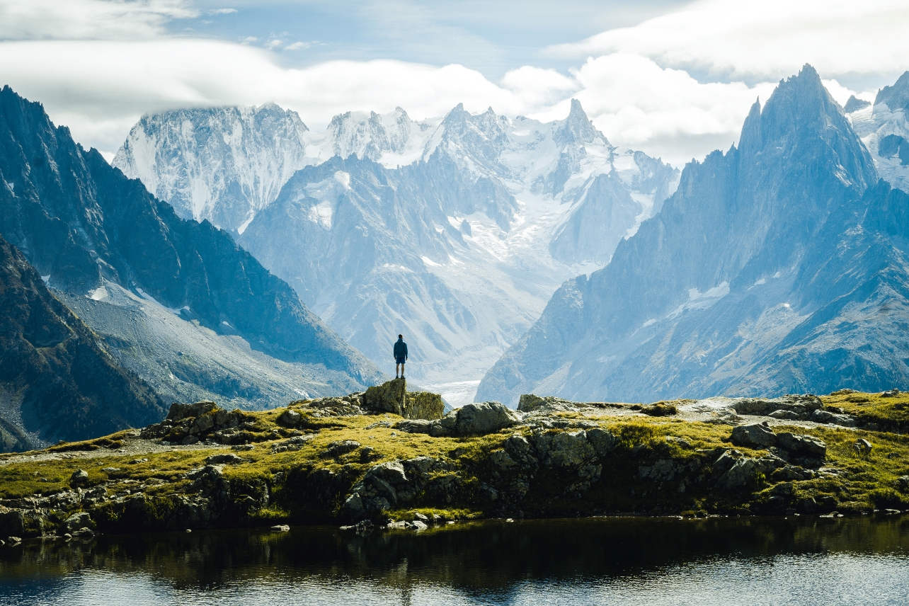 A person stands on a grassy hill, overlooking a serene lake and majestic snow-capped mountains under a cloudy sky.