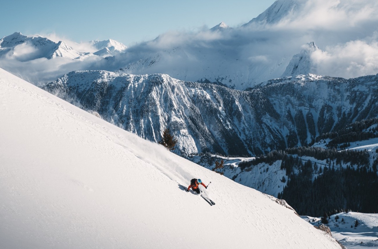 A skier in a red jacket descends a snowy mountain slope with towering, snow-covered peaks in the background under a blue sky with clouds.