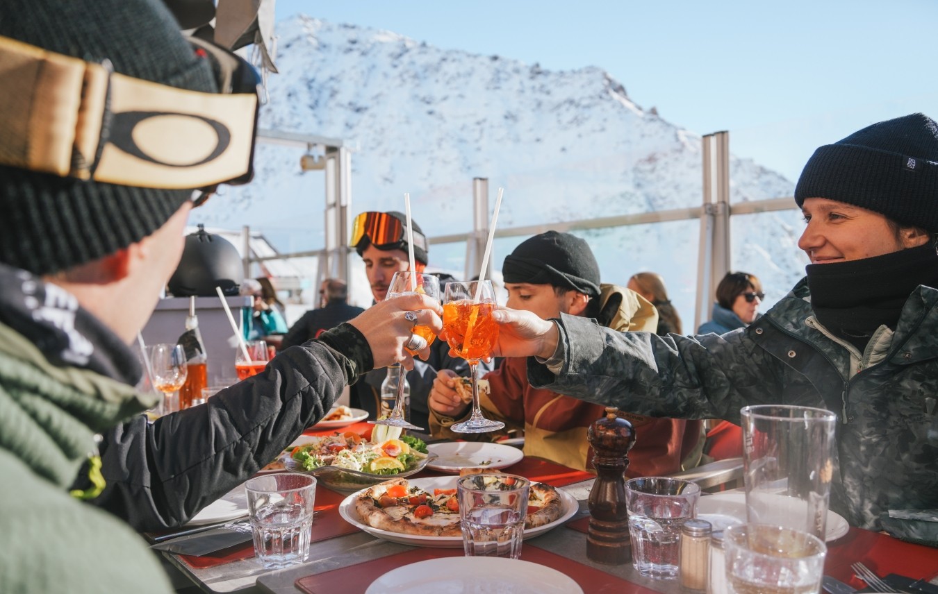 Sunset view of a ski rack filled with skis and snowboards near a railing, with people and mountains silhouetted in the background.