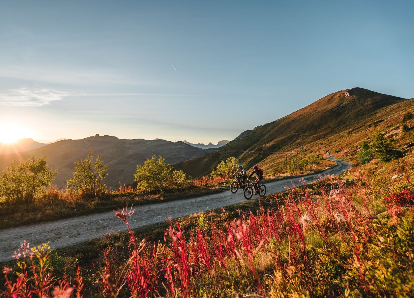 Two cyclists ride on a mountain trail at sunrise, surrounded by vibrant foliage and distant peaks under a clear sky.