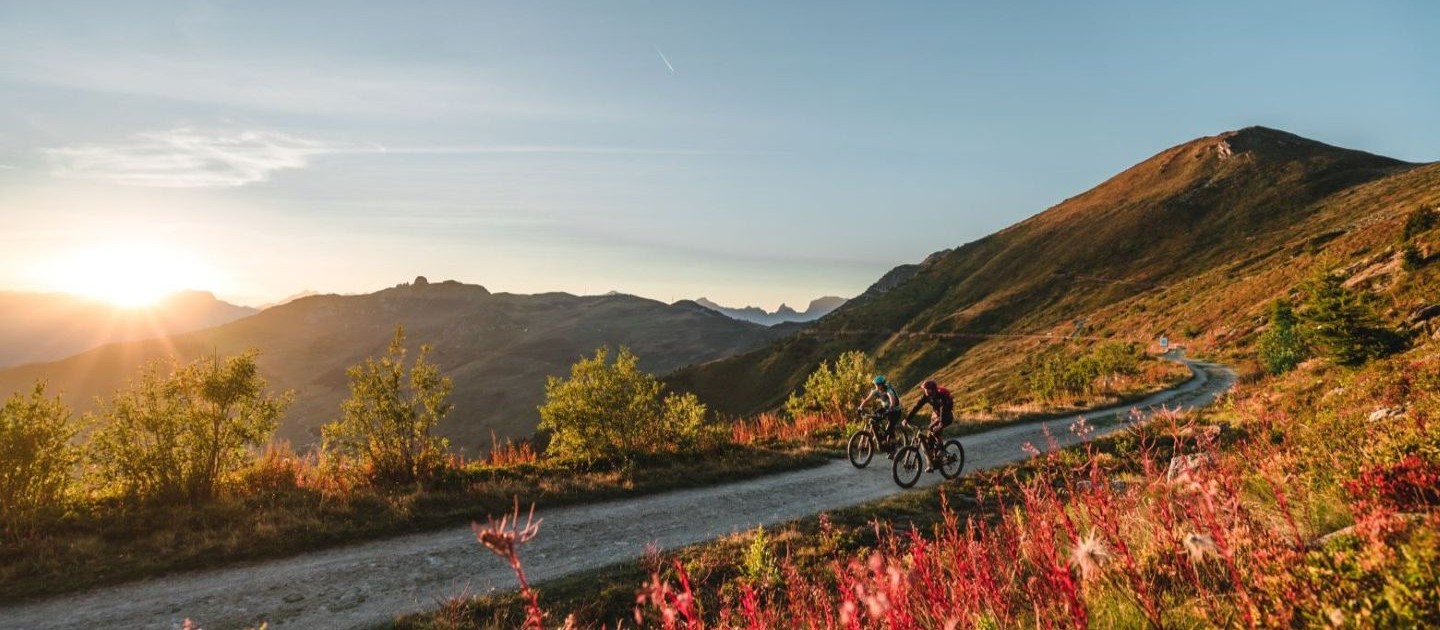 Verbier - summer biking Two cyclists ride on a mountain trail at sunrise, surrounded by vibrant foliage and distant peaks under a clear sky.
