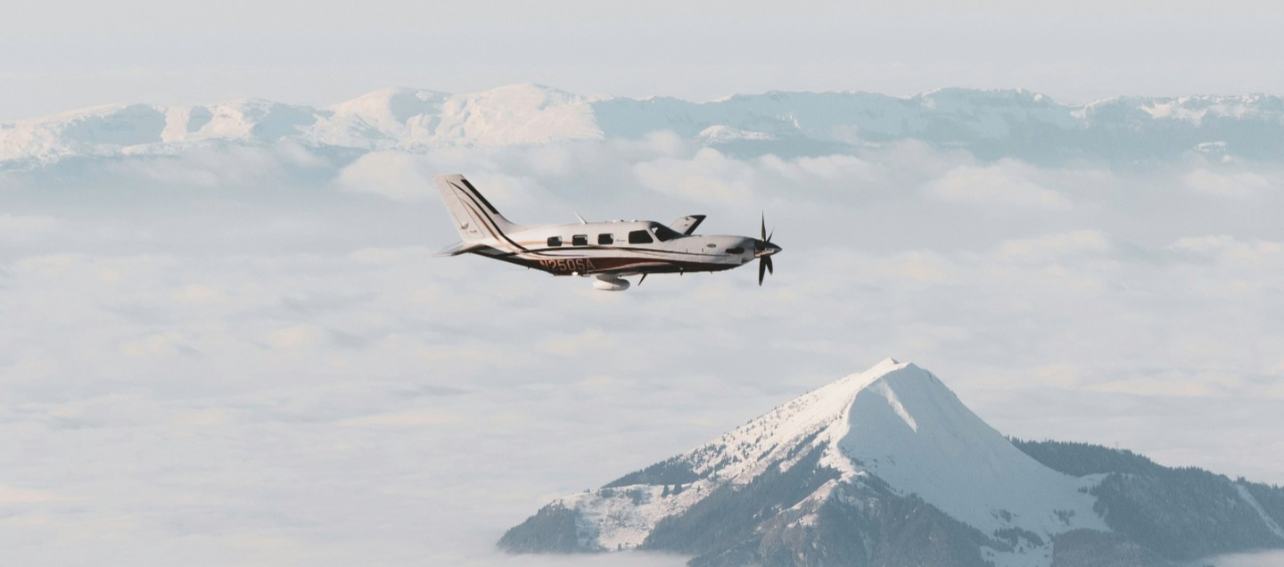 Small airplane flying above clouds with a snow-capped mountain peak visible below in the background.
