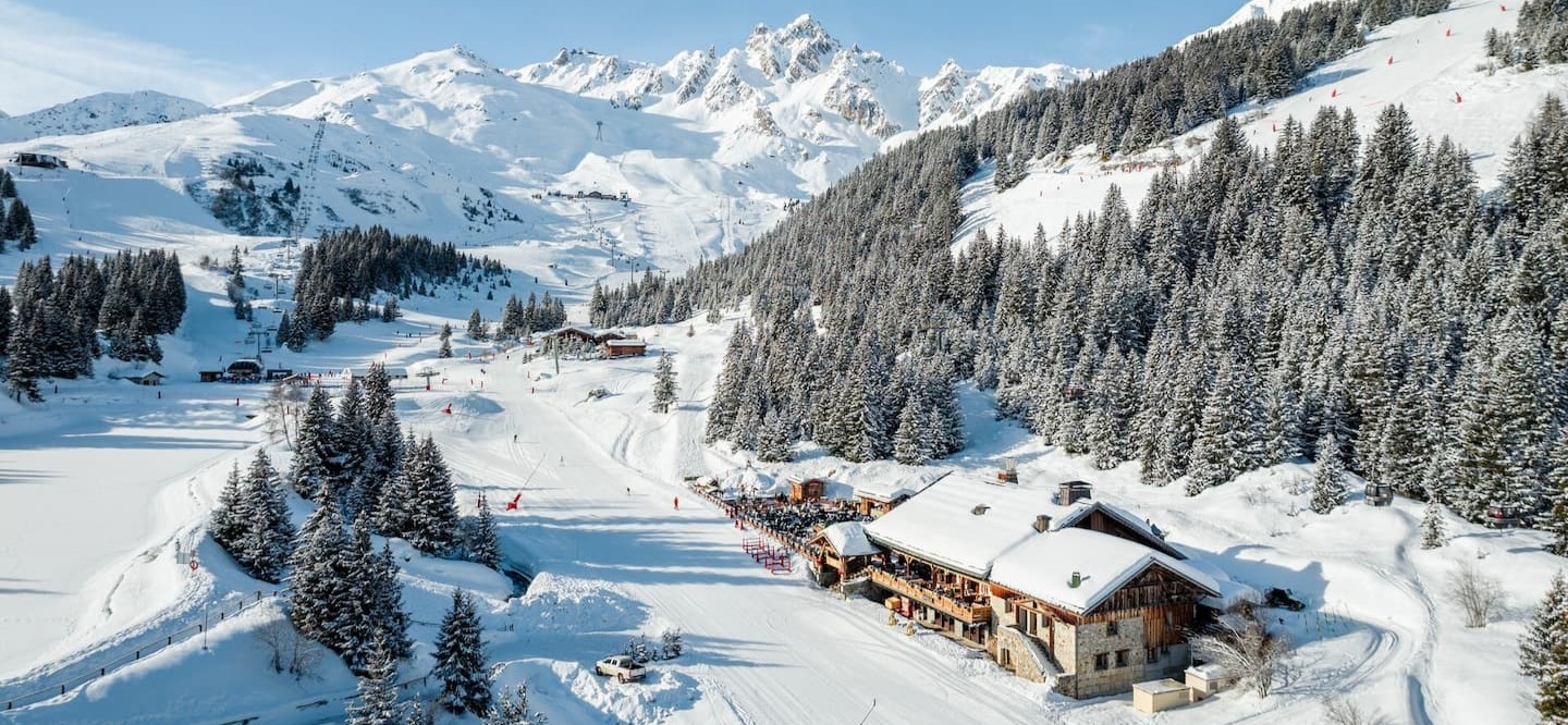 Snowy alpine scene with chalets, dense evergreen trees, and ski slopes surrounded by towering mountains under a clear blue sky.