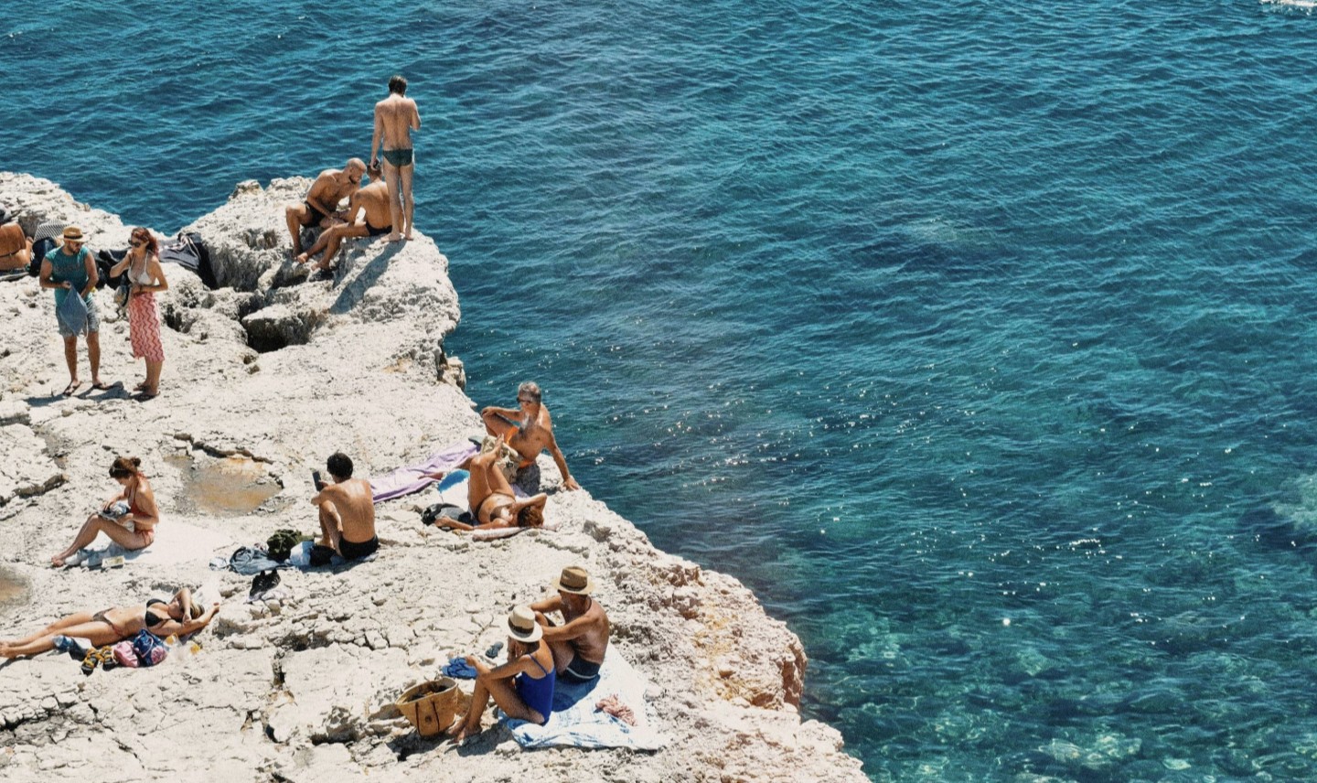 Boats and swimmers in a clear turquoise cove surrounded by rocky cliffs and green foliage under a bright blue sky.