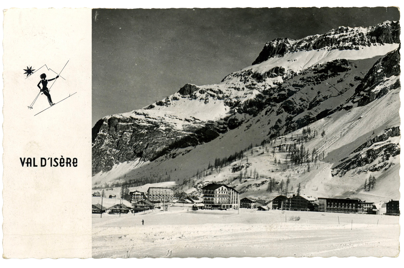 Vintage black-and-white postcard of Val d'Isère: alpine village and hotels beneath steep snow-covered mountains, skier silhouette at left.