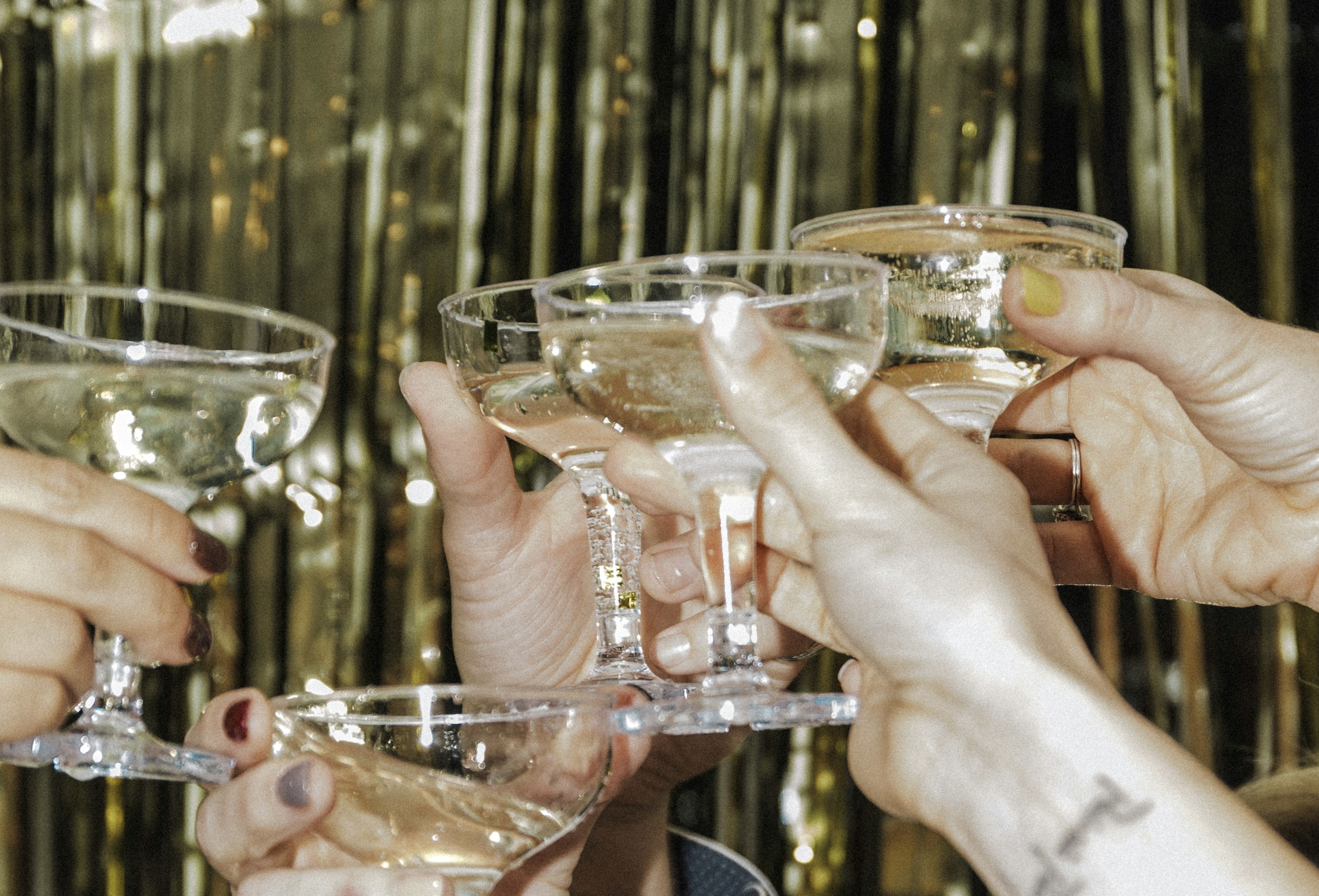 Hands holding champagne glasses in a celebratory toast against a gold tinsel backdrop.