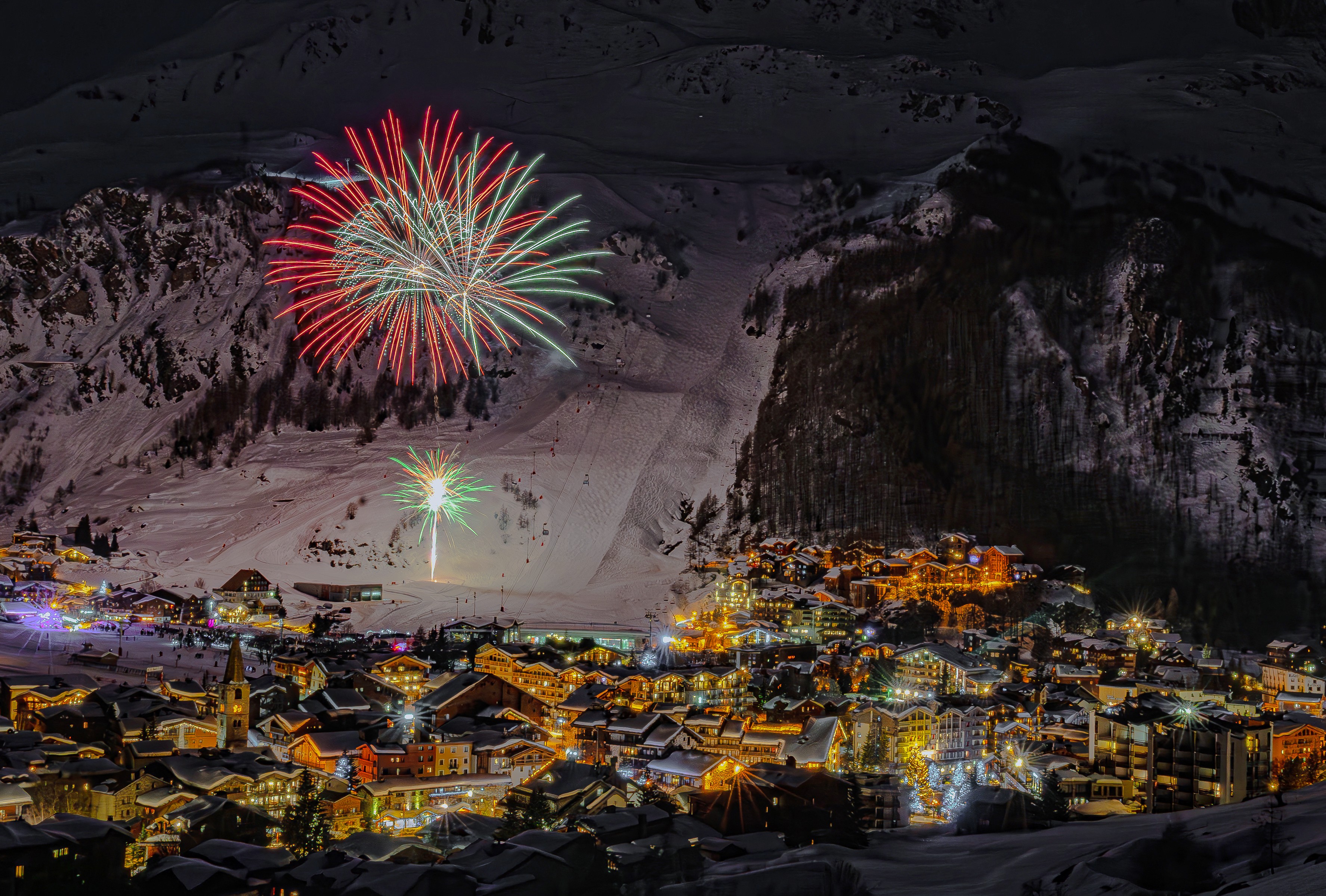 A snowy mountain village at night, illuminated by colorful fireworks in the sky and glowing lights reflecting off the snow-covered landscape.