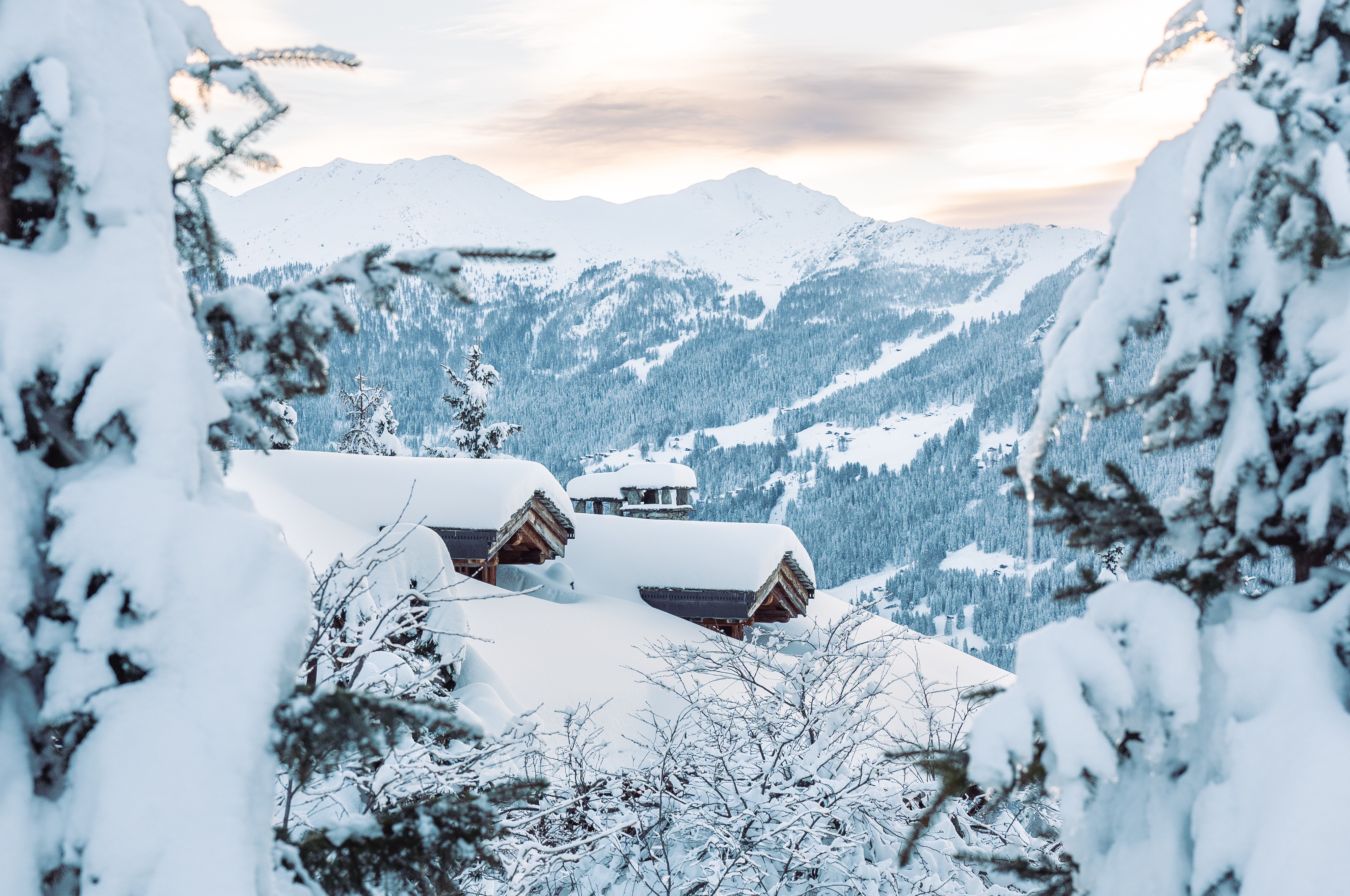 Snow-covered cabins nestled among trees with a backdrop of snowy mountains under a cloudy sky.