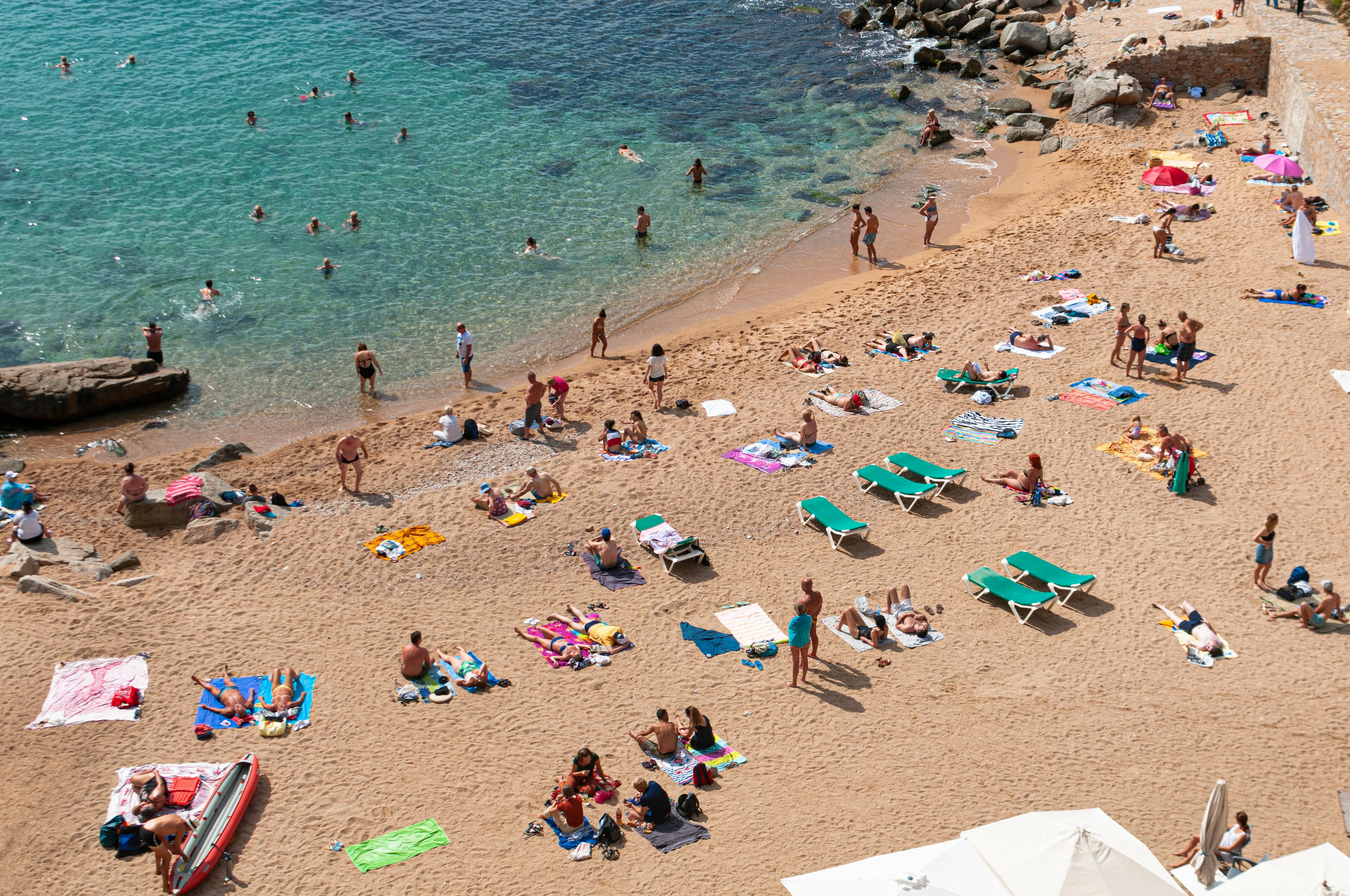 A crowded beach with sunbathers on towels and lounge chairs, and people swimming in the clear, blue water.