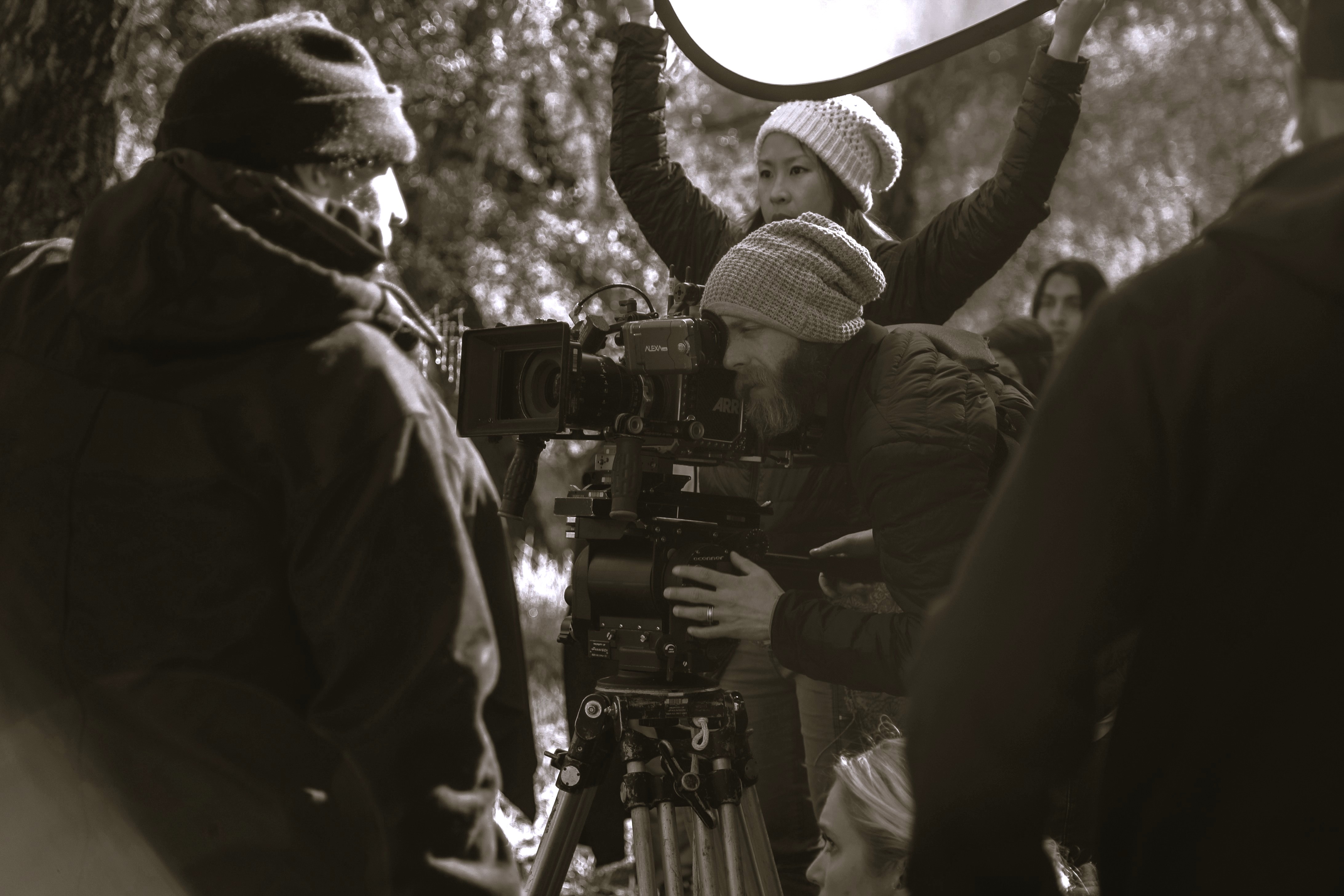 Film crew in outdoor setting; cameraman with beanie adjusts camera, woman holds reflector, others nearby, trees in background.