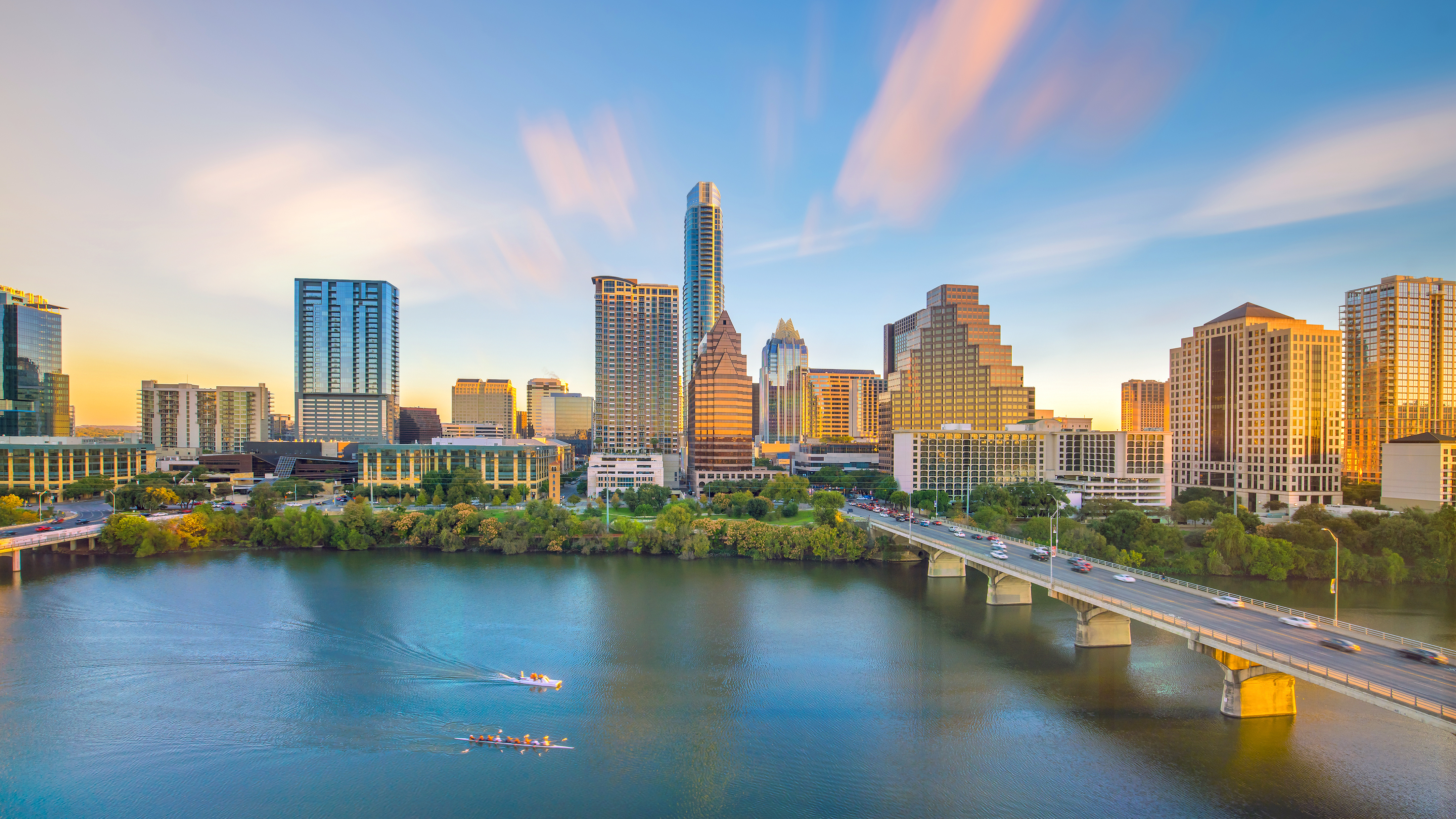 Skyline of a city with modern skyscrapers, a river with boats in the foreground, and a bridge to the right under a vibrant, partly cloudy sky.