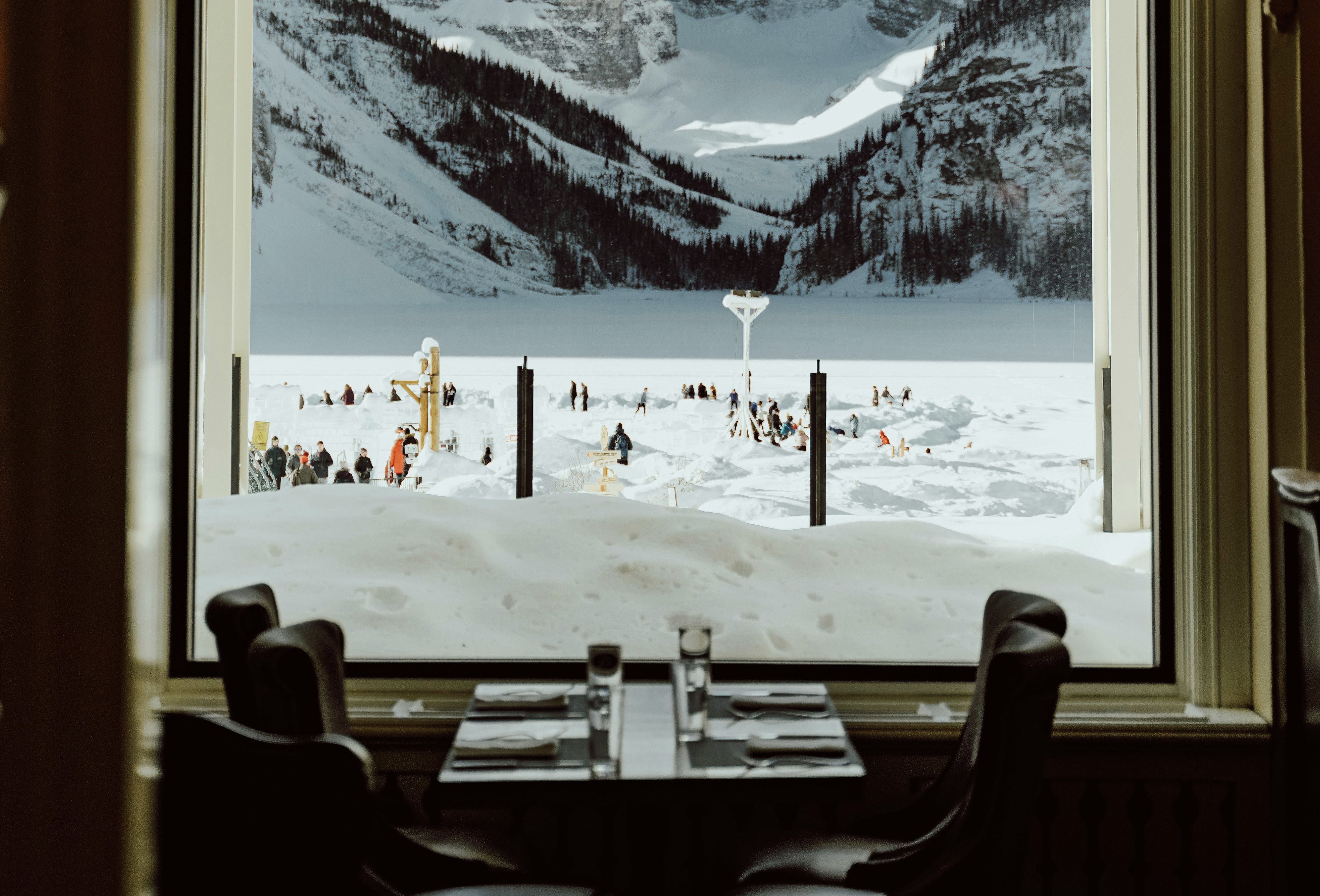 View from a cozy restaurant window showing a snowy landscape with people enjoying winter activities by a frozen lake and mountains in the background.