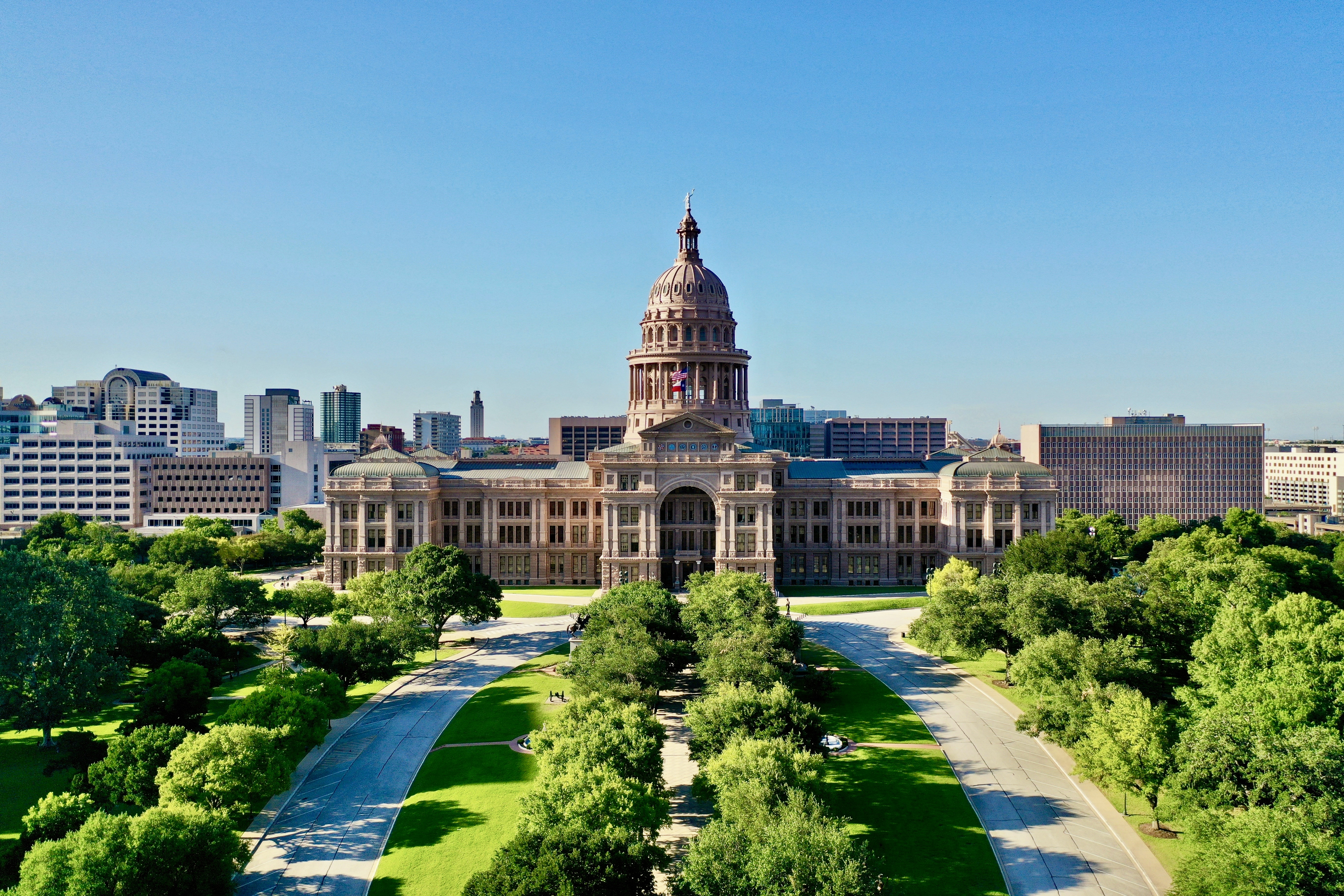 Aerial view of a large capitol building with a prominent dome, surrounded by greenery, set against a clear blue sky and a cityscape backdrop.
