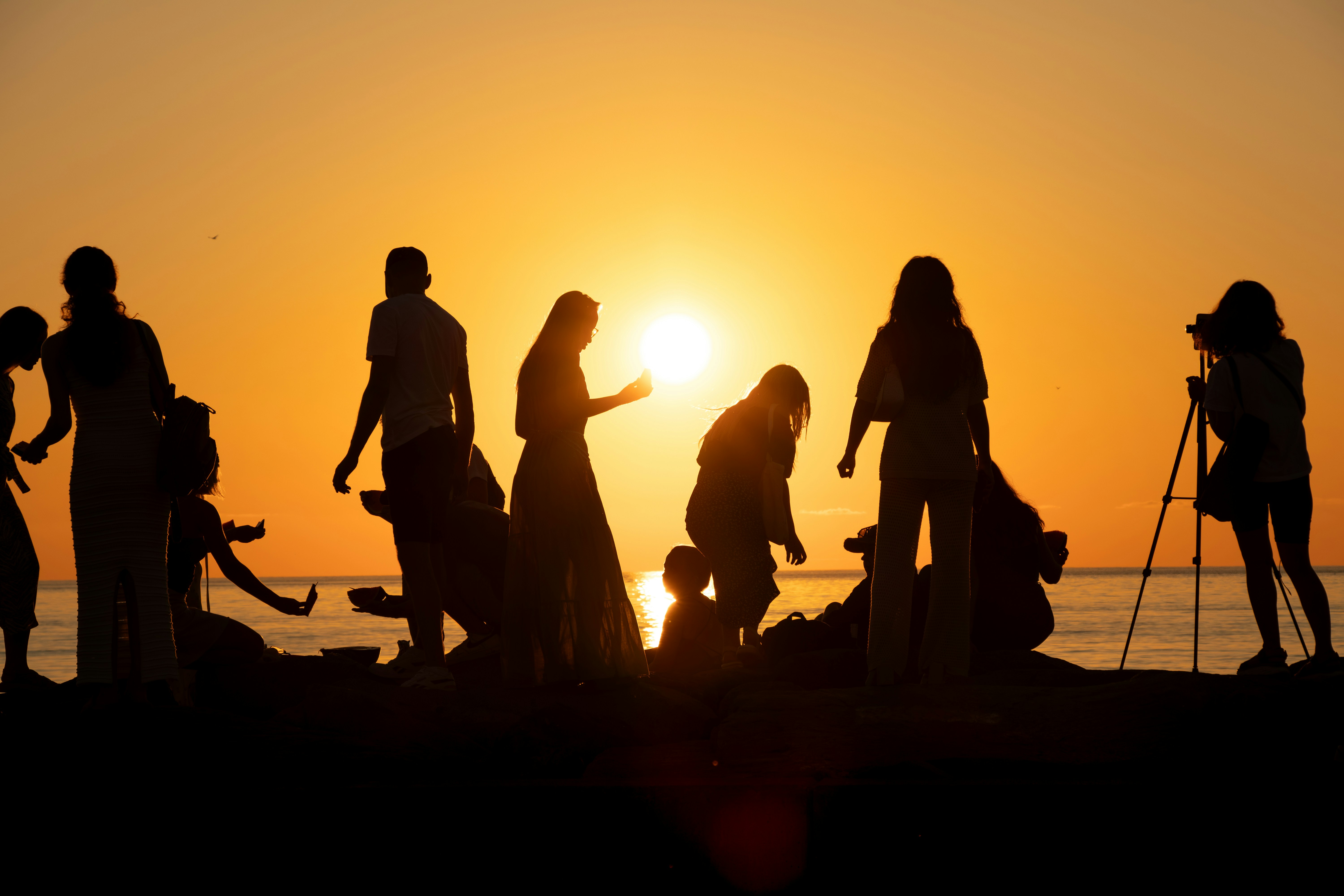 Silhouettes of people gathering at sunset near the ocean, with one person using a camera on a tripod.