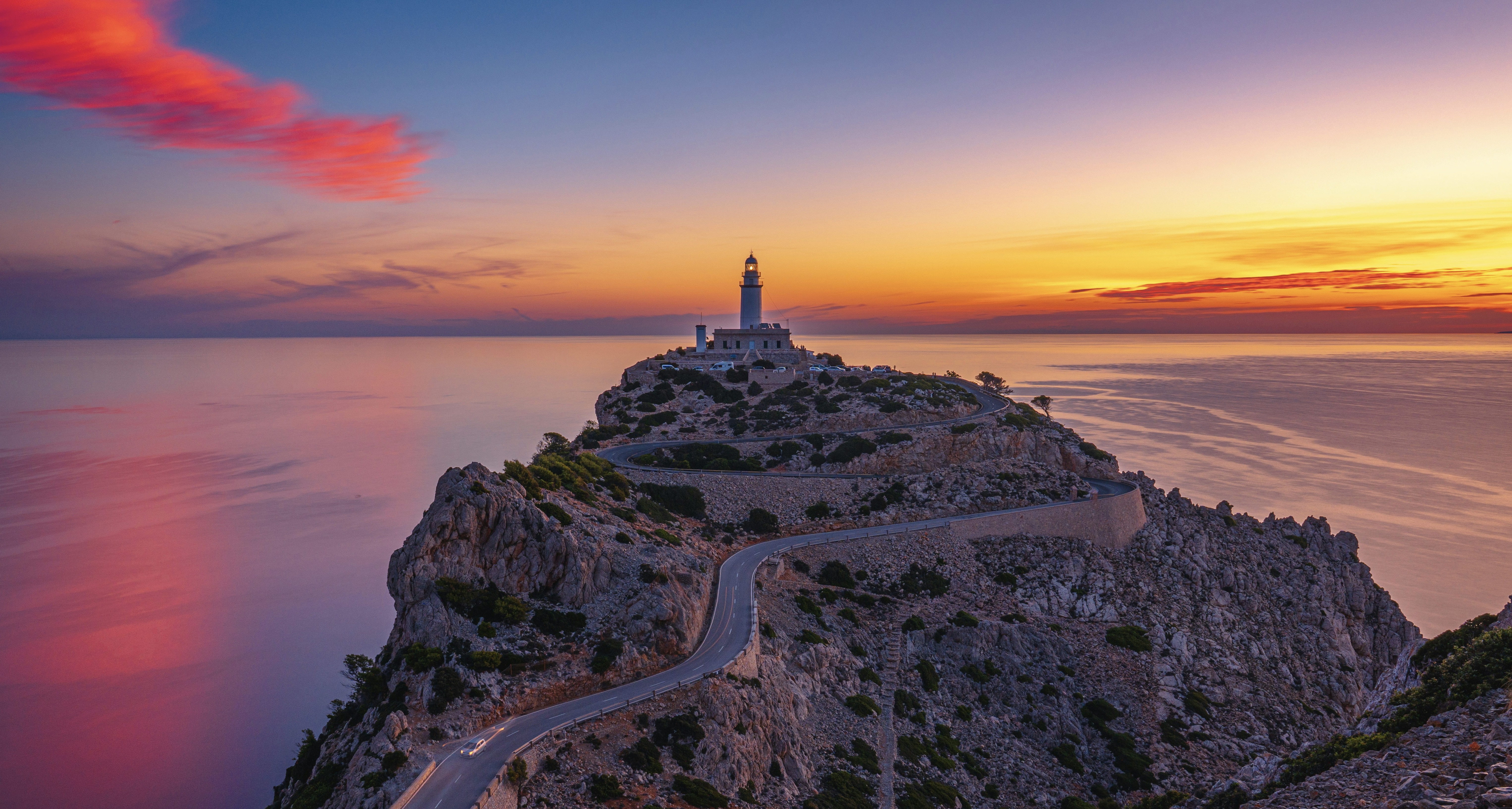 A winding road leads to a lighthouse atop a rocky cliff at sunset, with vibrant skies reflecting on calm ocean waters.