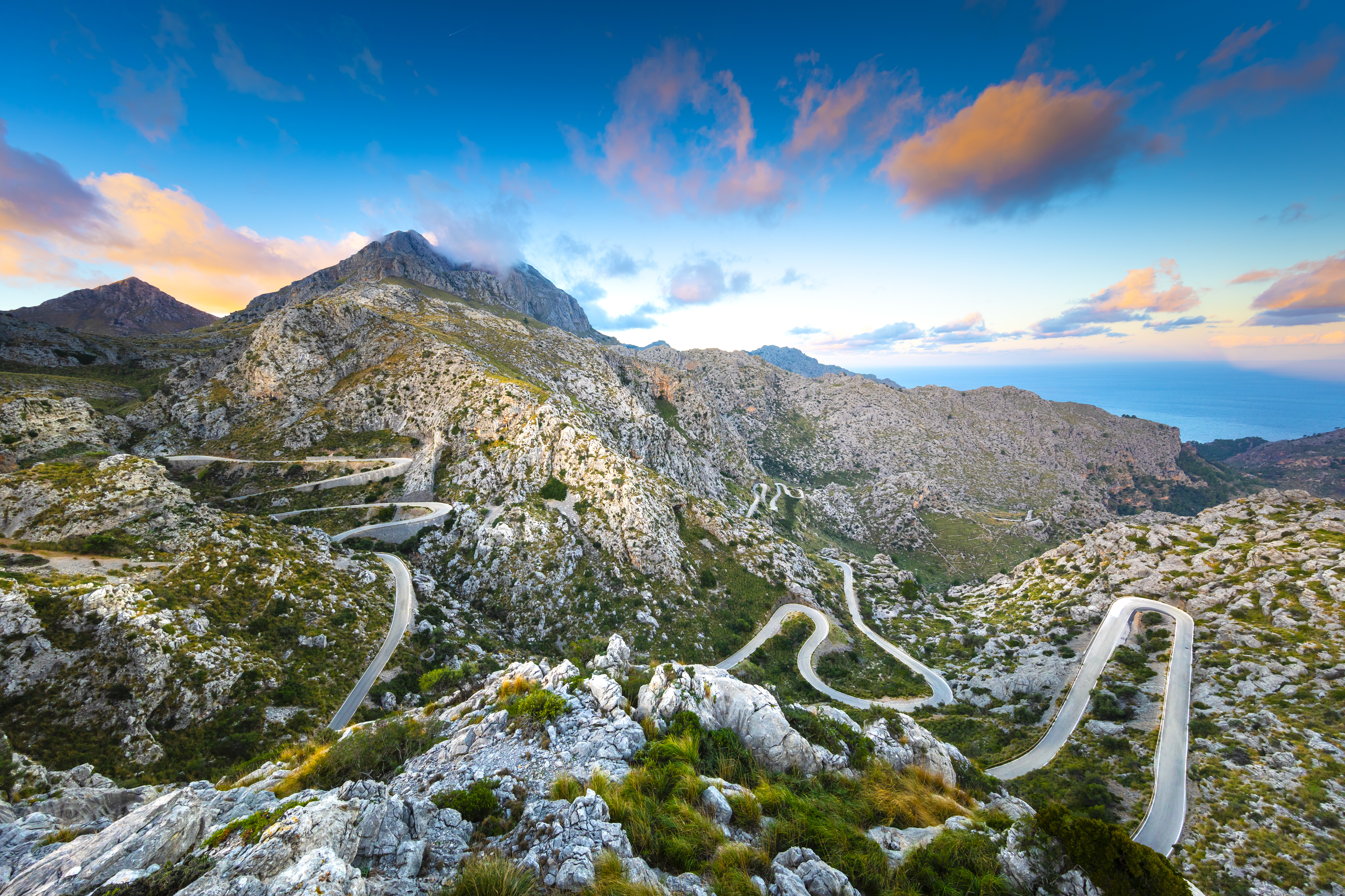 Winding mountain road with sharp curves, surrounded by rocky terrain and under a vibrant sky with scattered clouds.
