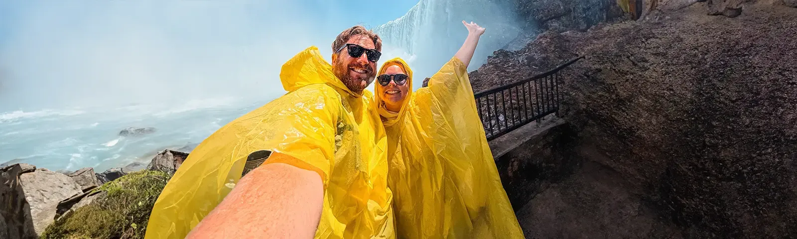 Two people in yellow rain ponchos take a selfie near a misty waterfall, both smiling with one raising an arm, rocky path beside them.