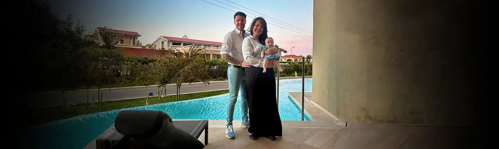 A couple stands by a pool, smiling, with the woman holding a baby. Houses and a pink sky are visible in the background.