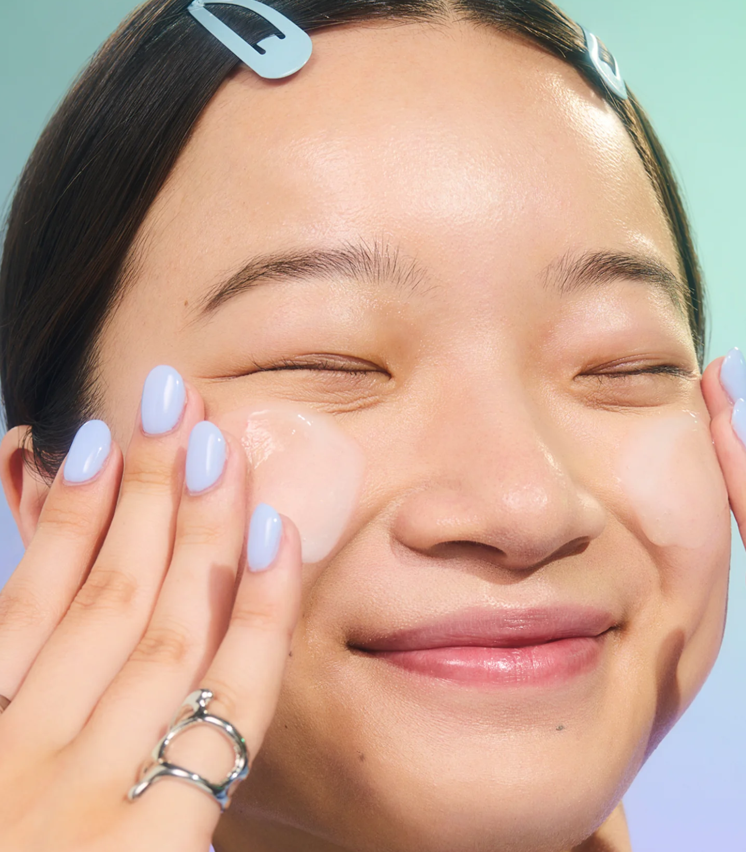 Person with closed eyes applying cream to their cheeks, smiling, with hair clips and pastel blue nail polish.
