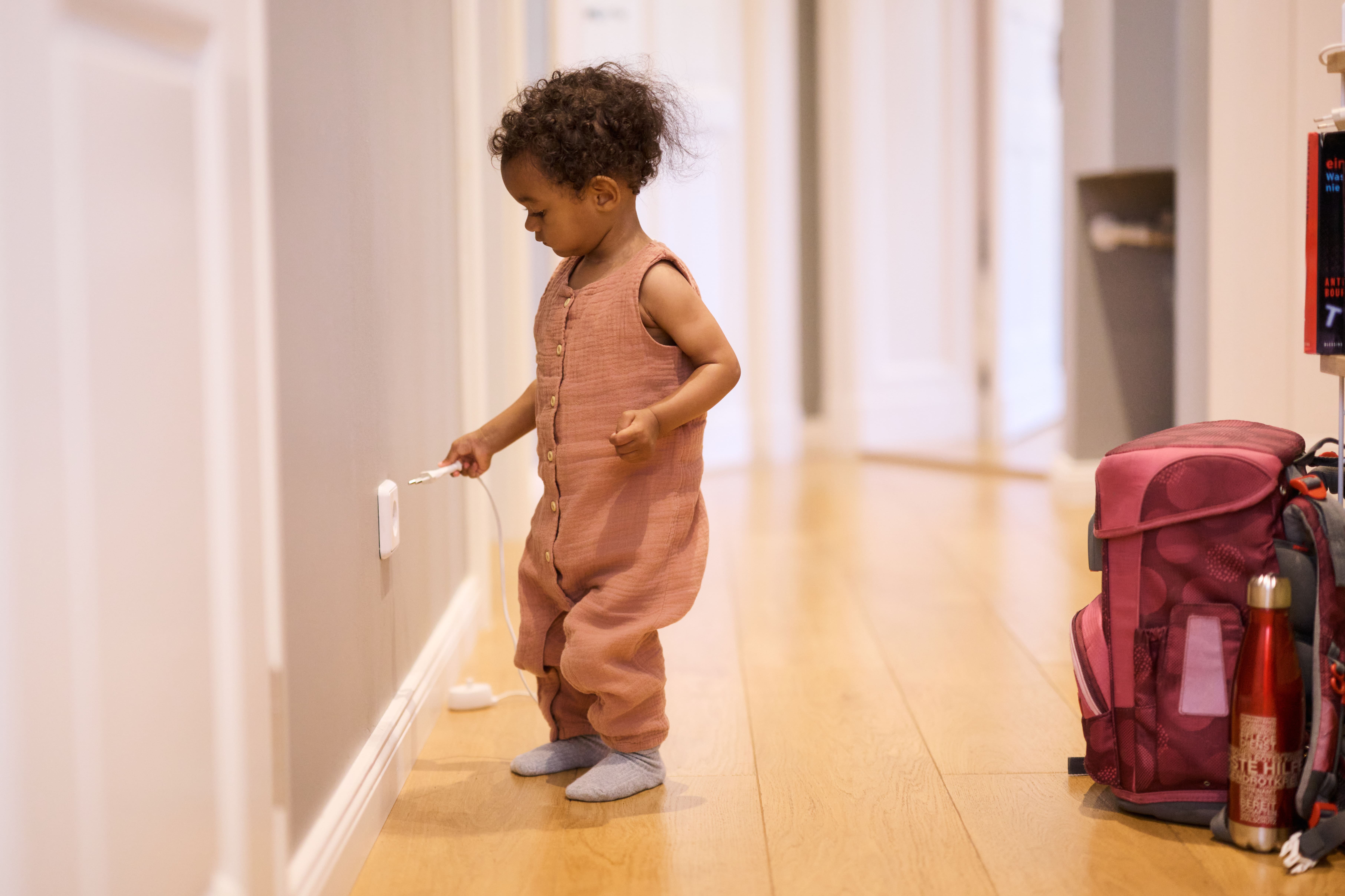 A toddler in a pink outfit stands in a hallway, reaching for an electrical outlet. A red backpack is nearby on the wooden floor.